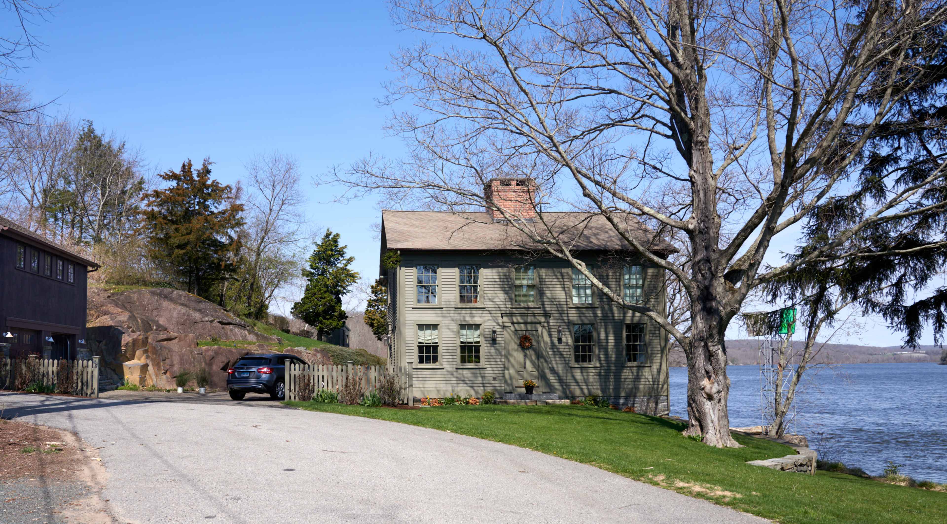 A two-story wooden house with a sloped roof is situated near a body of water, surrounded by sparse trees and a gravel road.