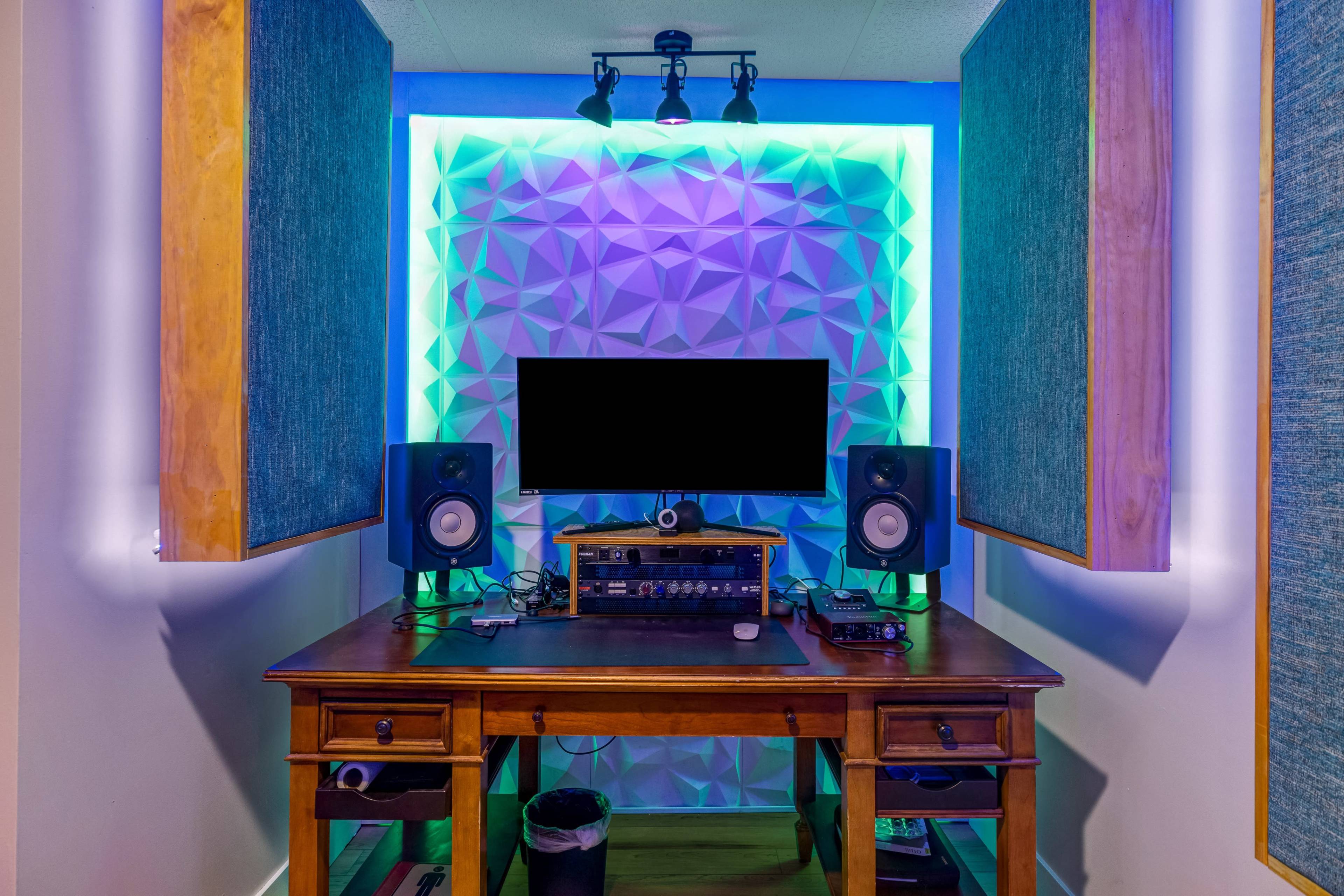 A home recording studio with a wooden desk, speakers on either side, and a monitor against a geometric-patterned wall lit with colored lights.