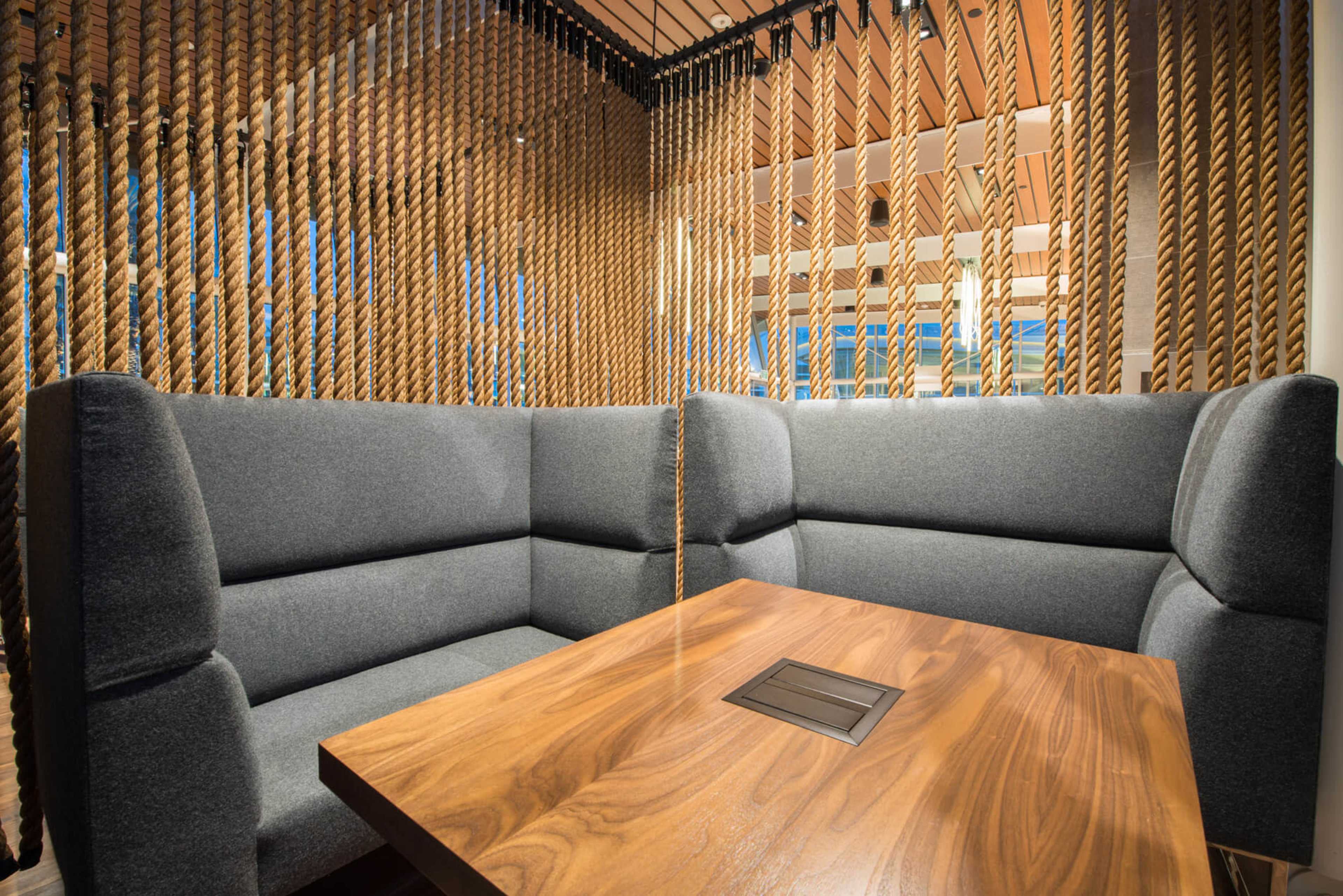 A modern, semi-enclosed seating area with gray upholstered benches and a wooden table surrounded by hanging ropes.