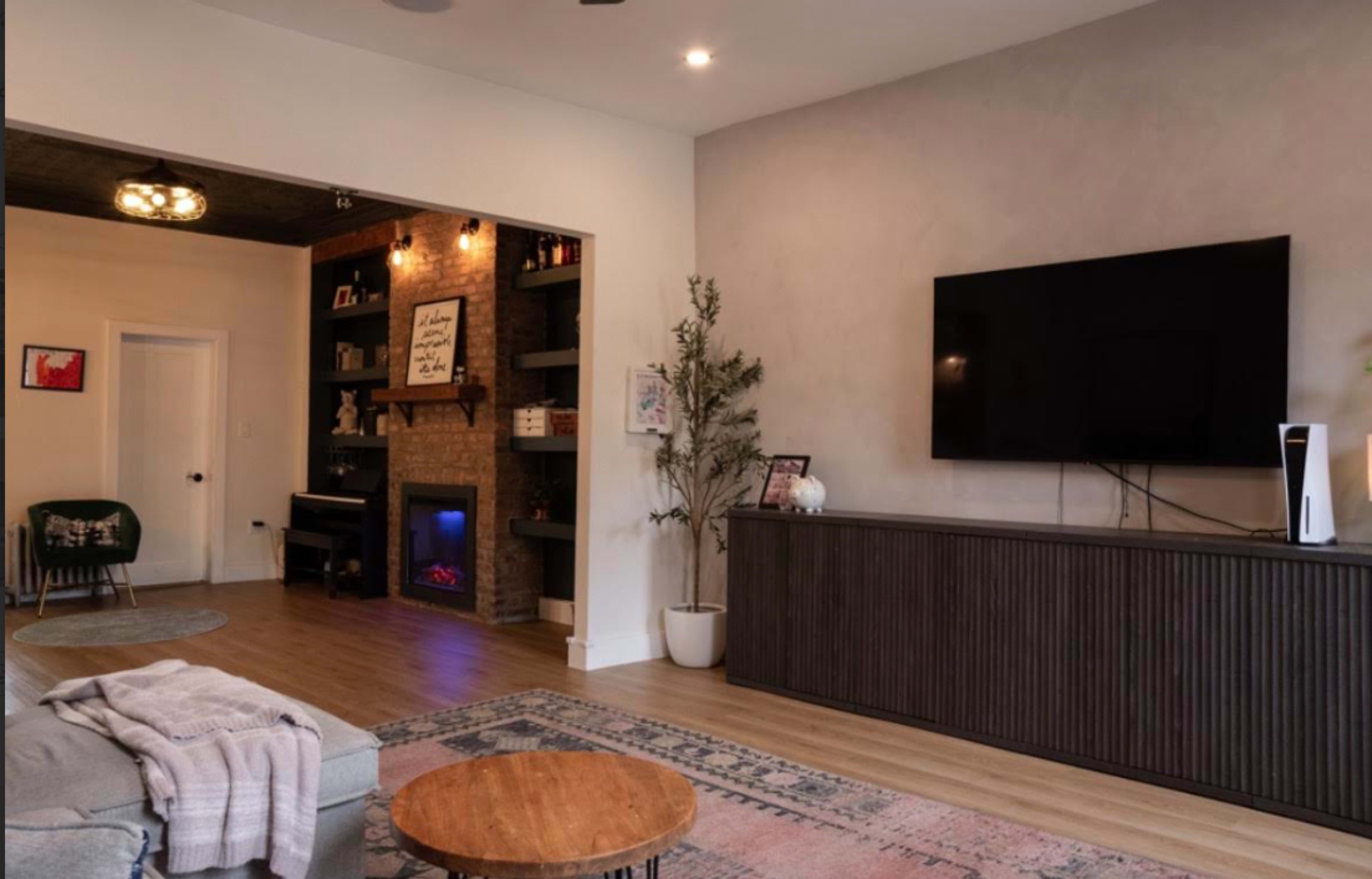 The image shows a modern living room featuring a wall-mounted television, a brick fireplace with an electric insert, and a cozy seating area with a coffee table.