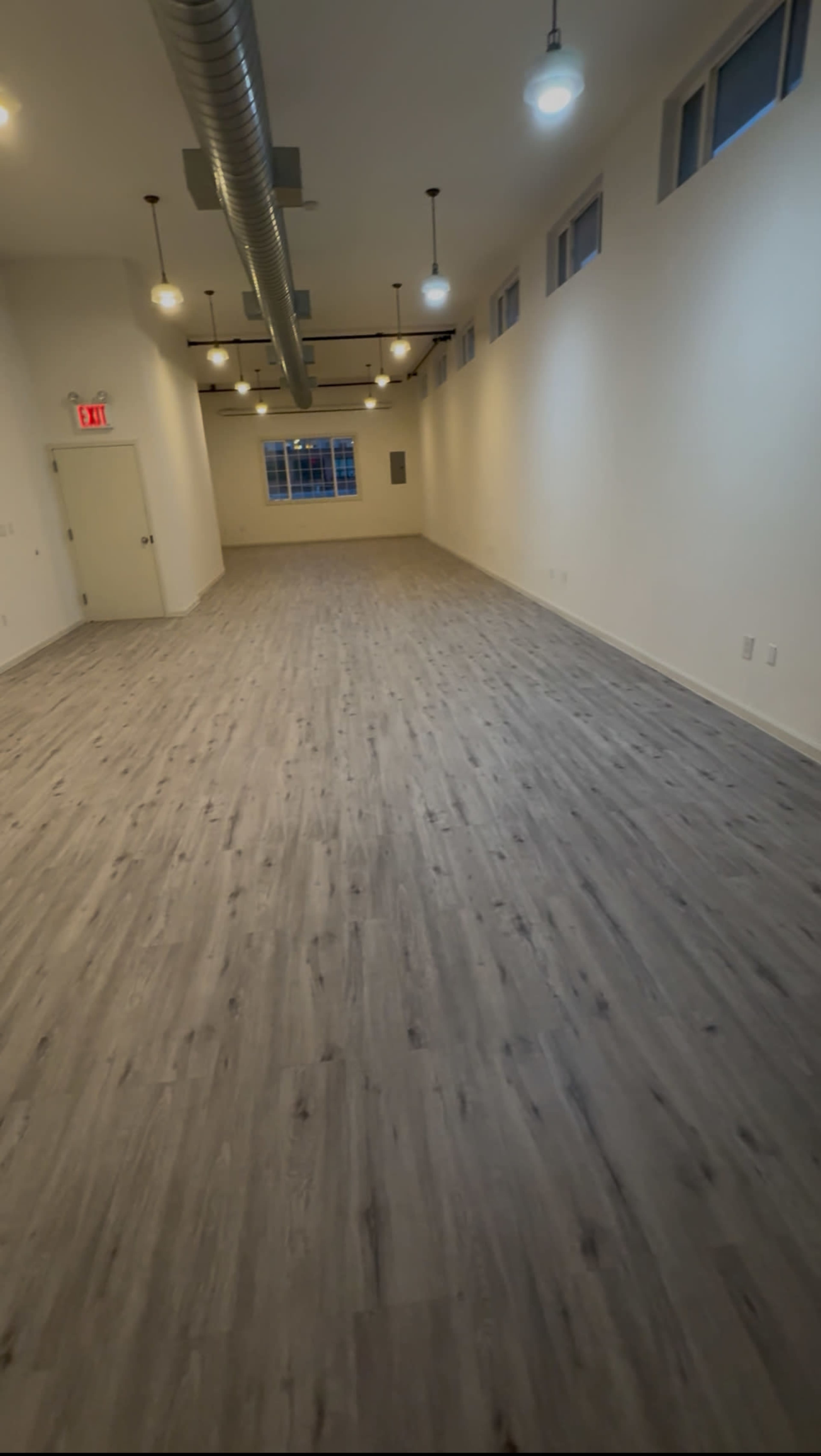 The image shows an empty, well-lit interior space with wooden flooring and large windows along one wall.