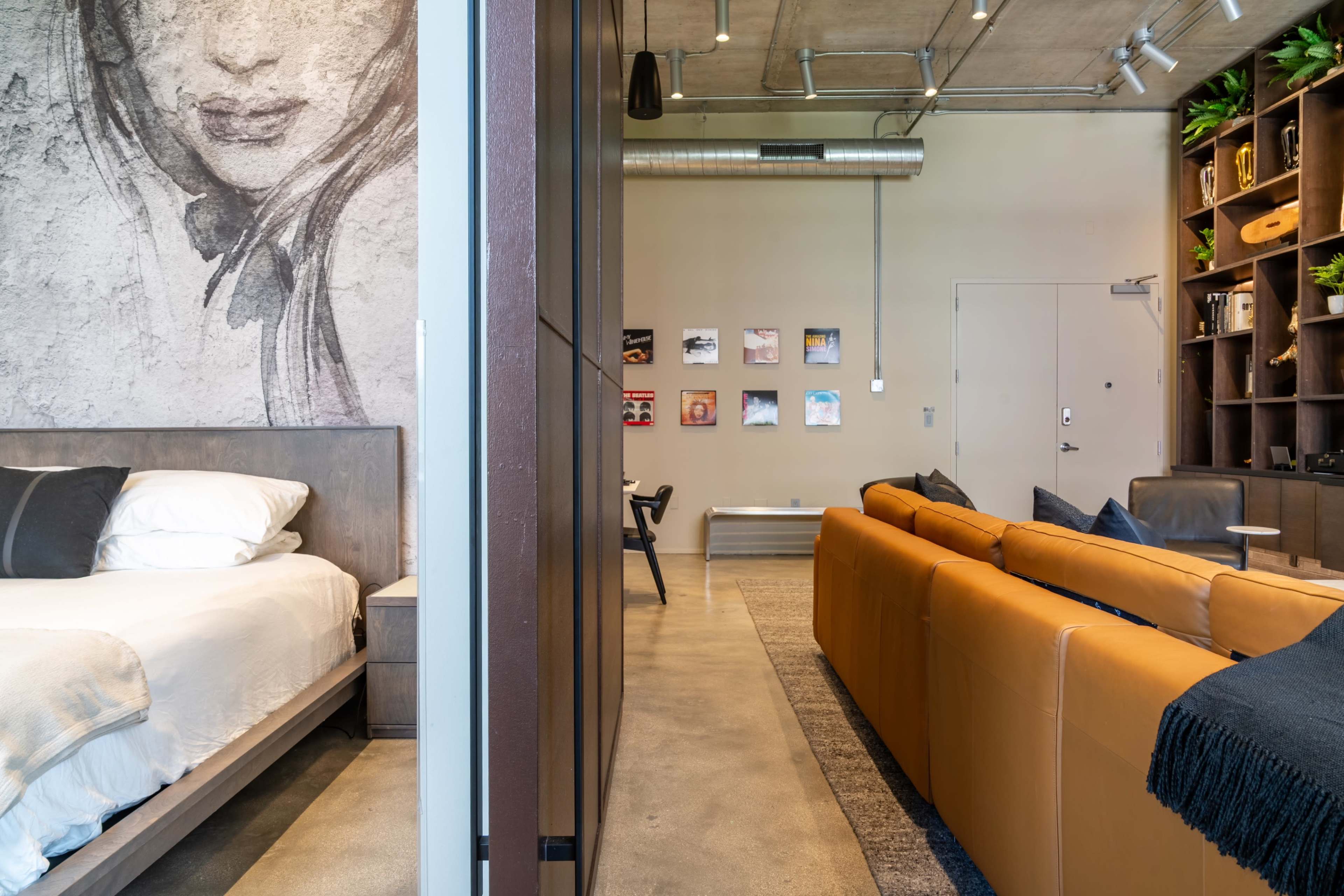 The image shows a modern interior space with a bedroom featuring a large artwork on the wall and a small living area with a leather sofa and a bookshelf.