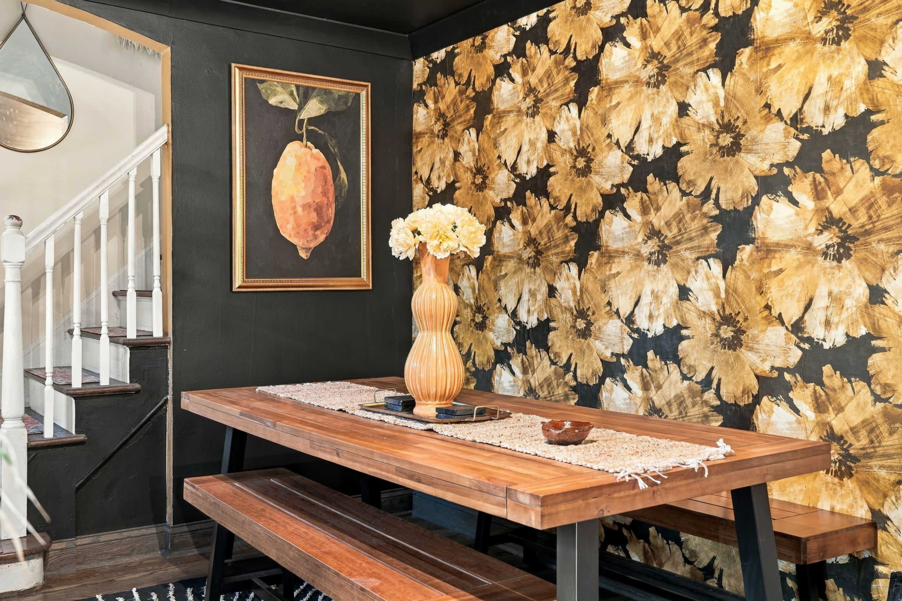 A dining area features a wooden table, a tall vase with flowers, and a wall adorned with large floral-patterned wallpaper and a framed painting of a lemon.