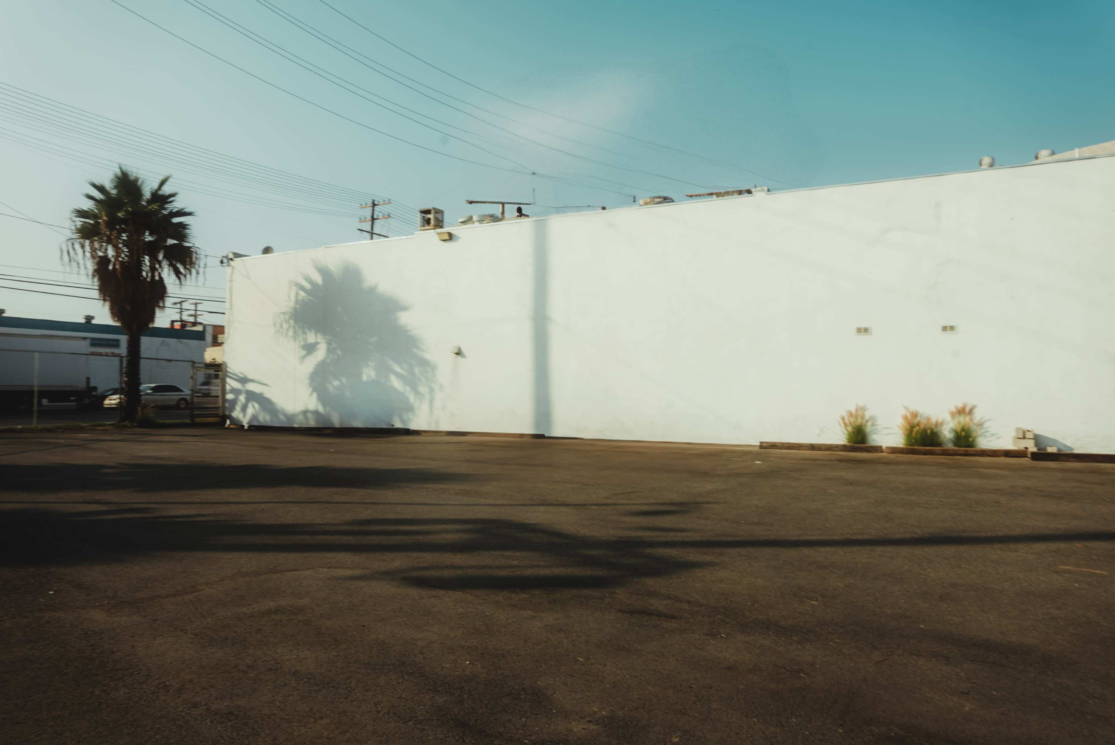 A large white wall casts a shadow on an empty asphalt parking lot under a clear blue sky.