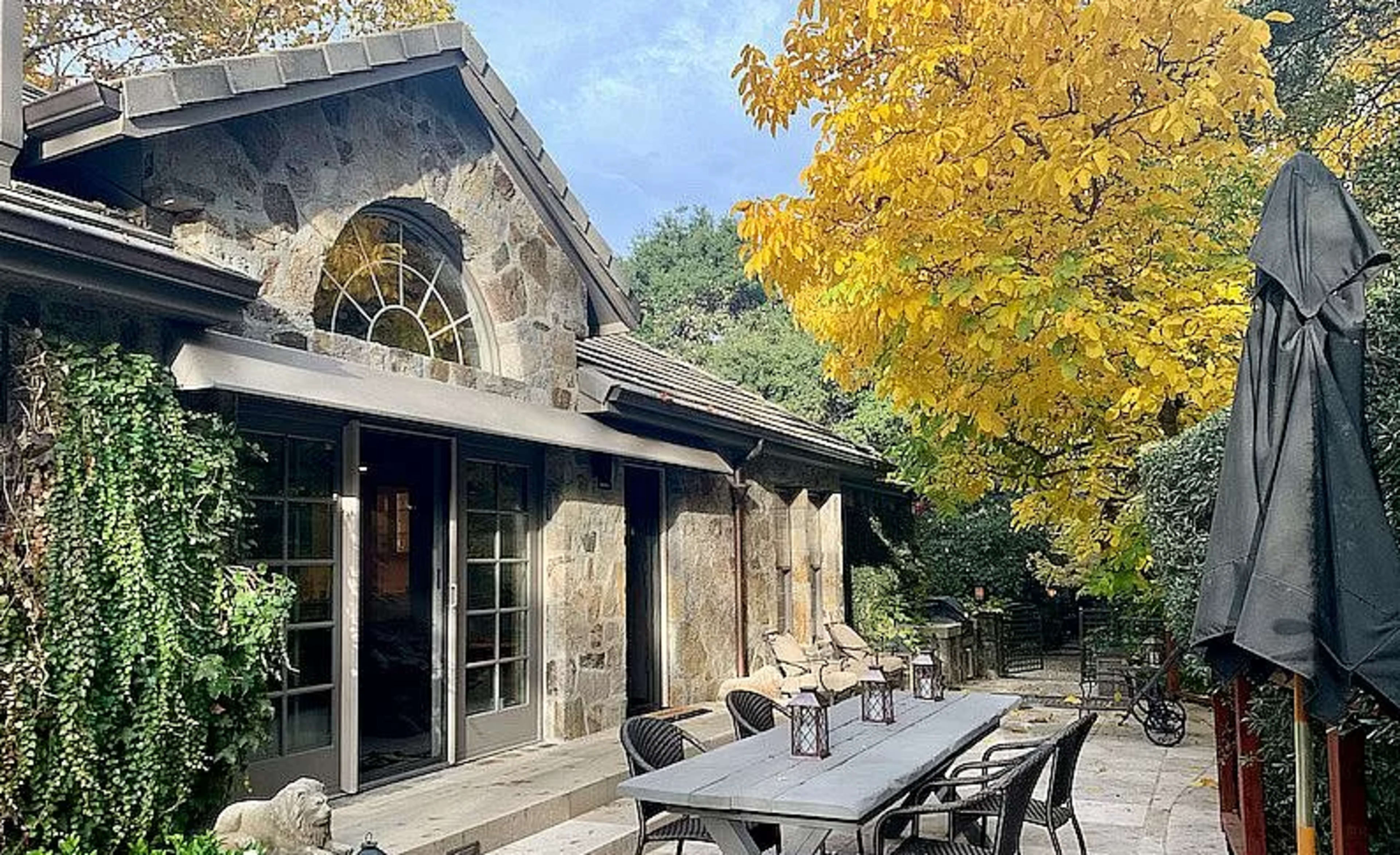 A stone house with large windows, a shaded outdoor dining area featuring a long table and chairs, and a vibrant yellow tree nearby.