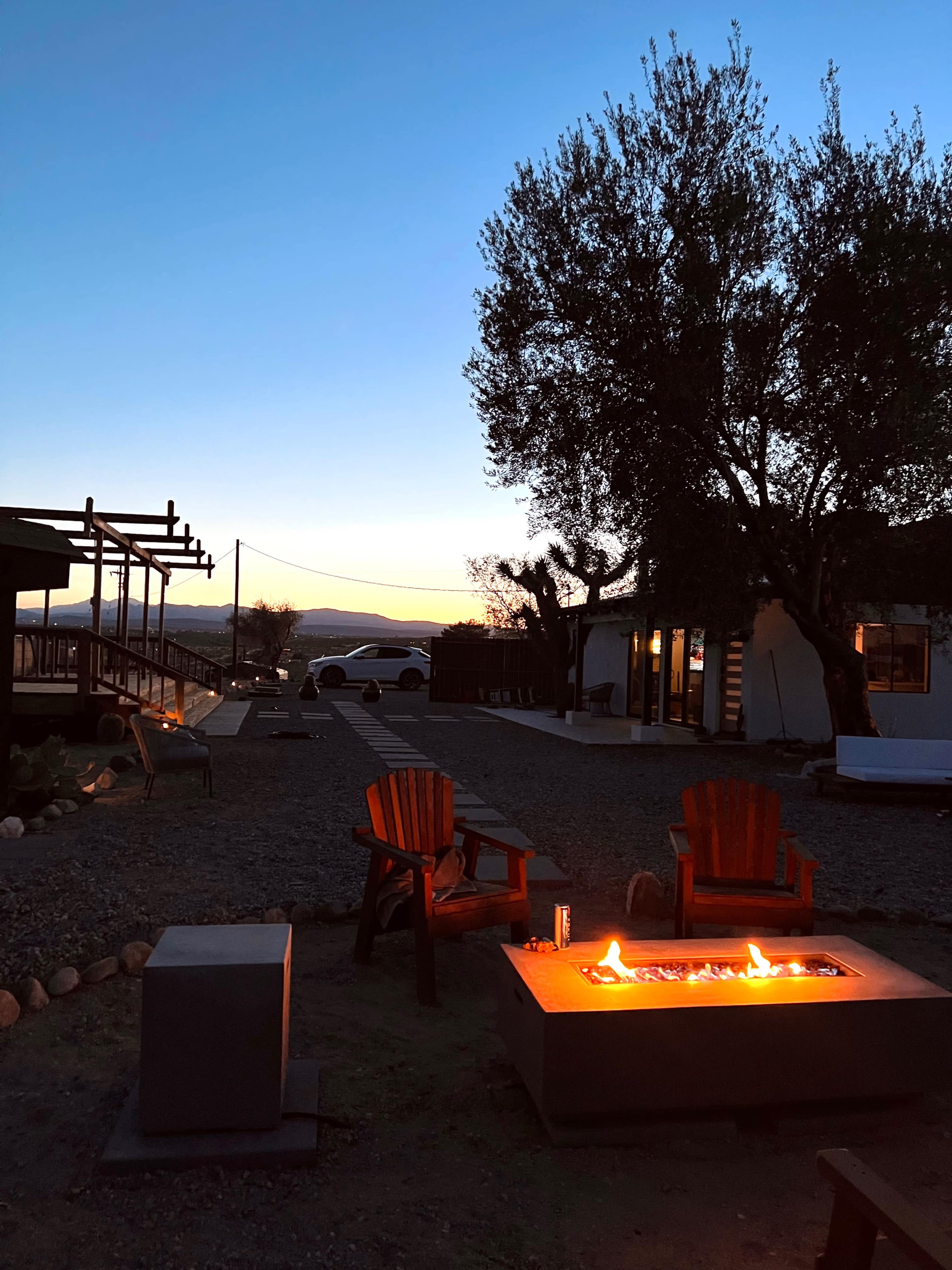 The image shows a gravel yard with a fire pit and Adirondack chairs, framed by a tree and a house against a twilight sky.
