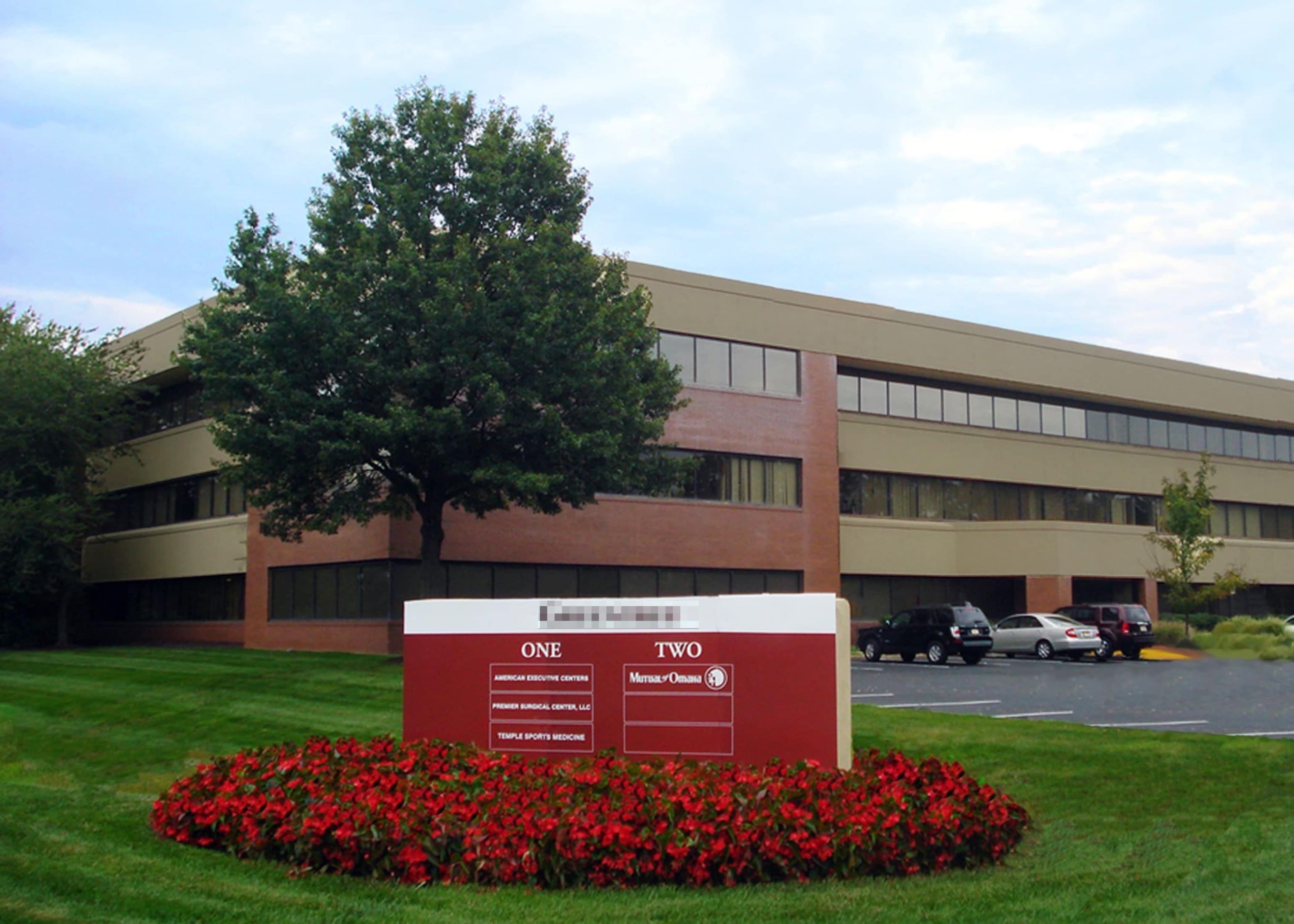 The image shows a three-story office building with a landscaped entrance featuring flower beds and a prominent sign indicating the building's name and number.