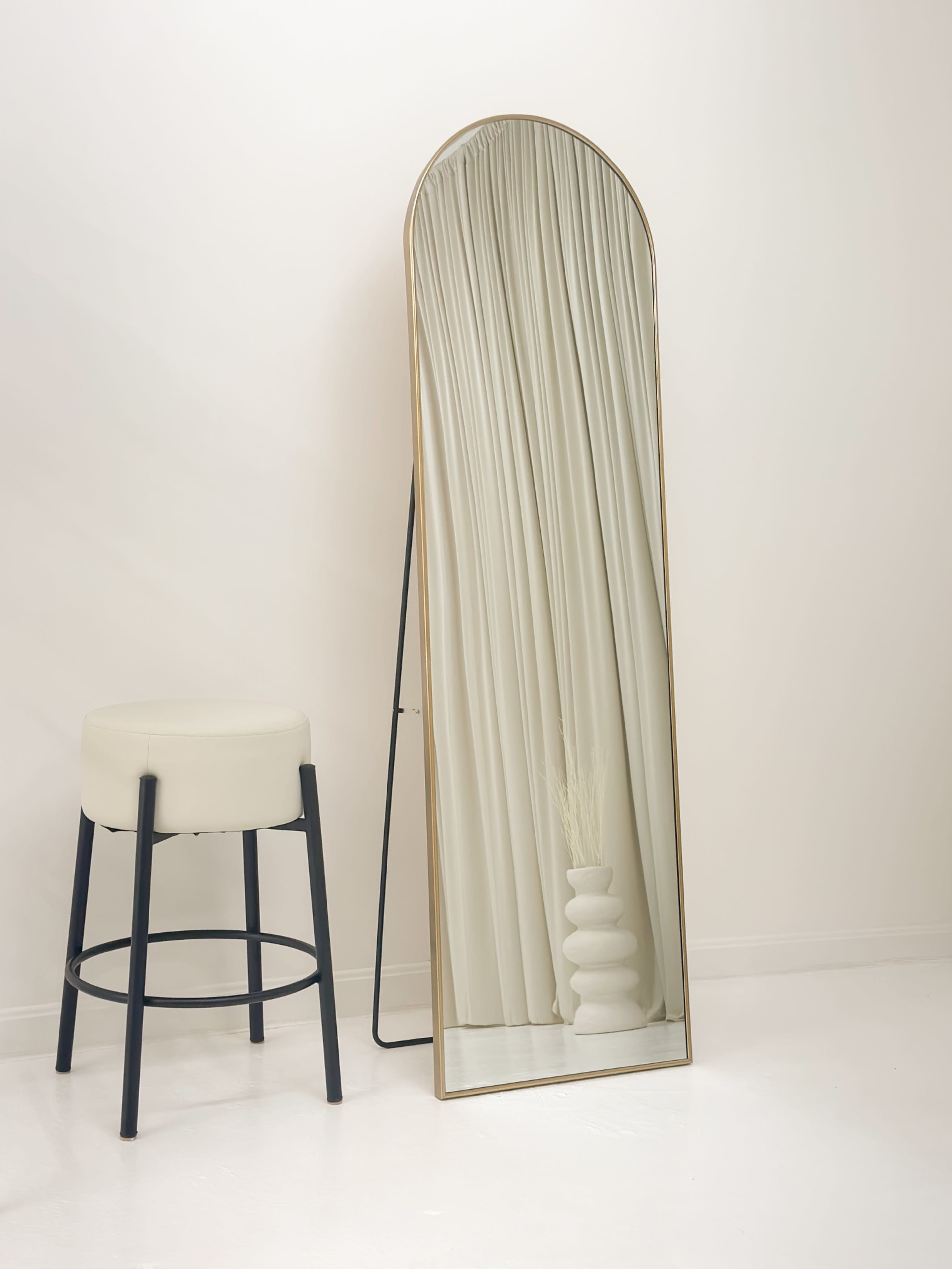 A tall mirror with an arched top stands against a wall next to a minimalist stool and a white vase on the floor.