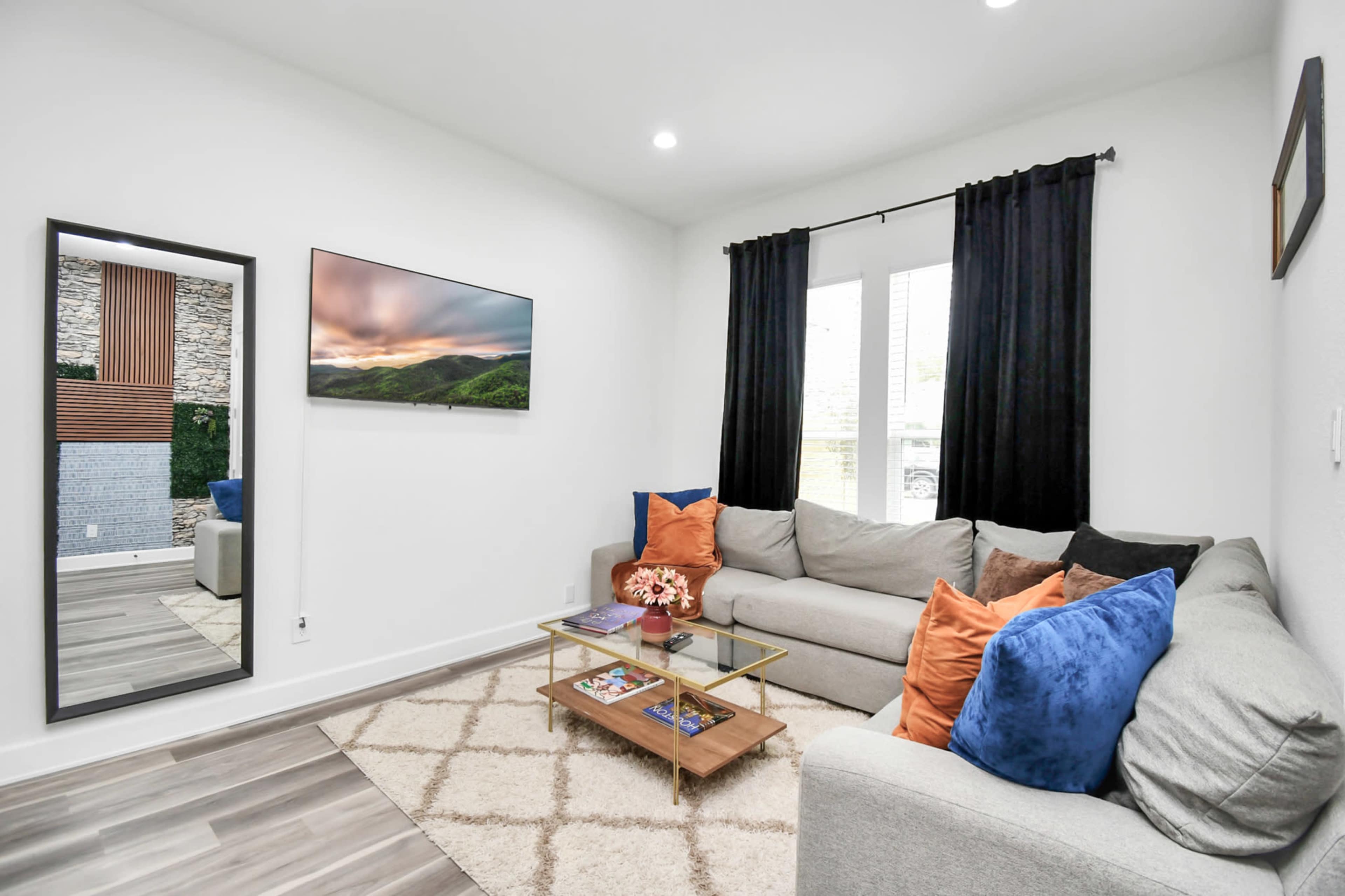 The living room features a gray sectional sofa, a glass coffee table with magazines, a large mirror, and a wall-mounted television above a decorative stone feature.