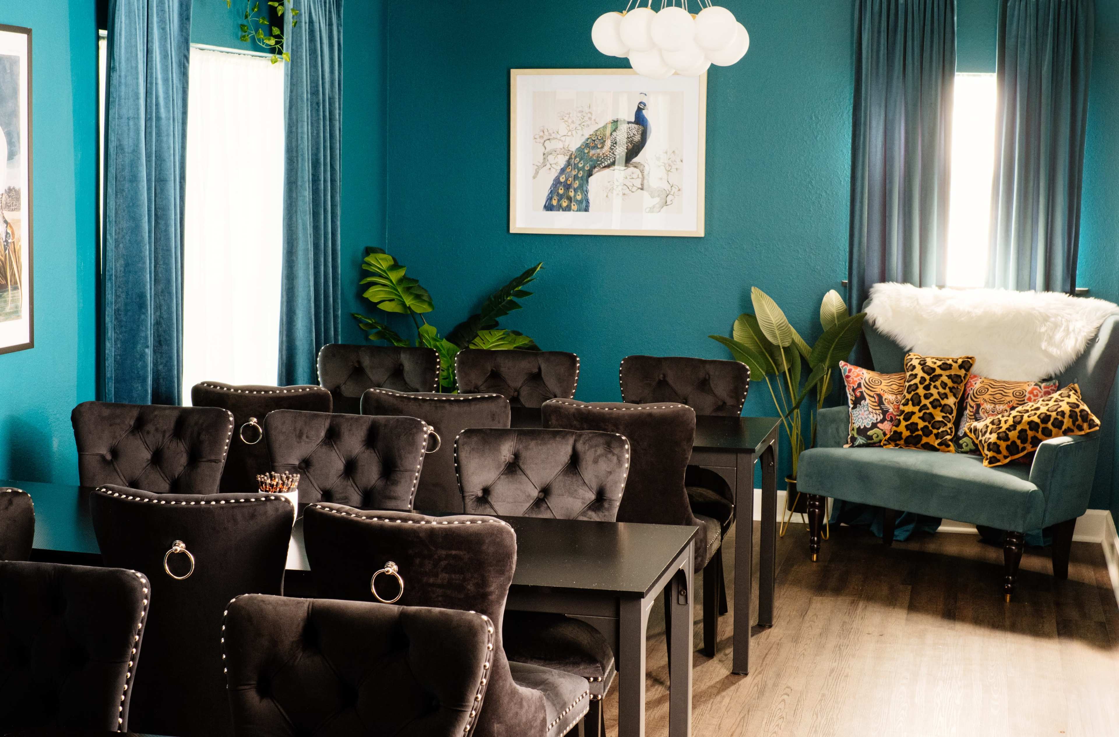 The image features a vibrant room with teal walls, decorated with a peacock print and equipped with several black velvet chairs around black tables, along with a cozy blue armchair adorned with decorative pillows.