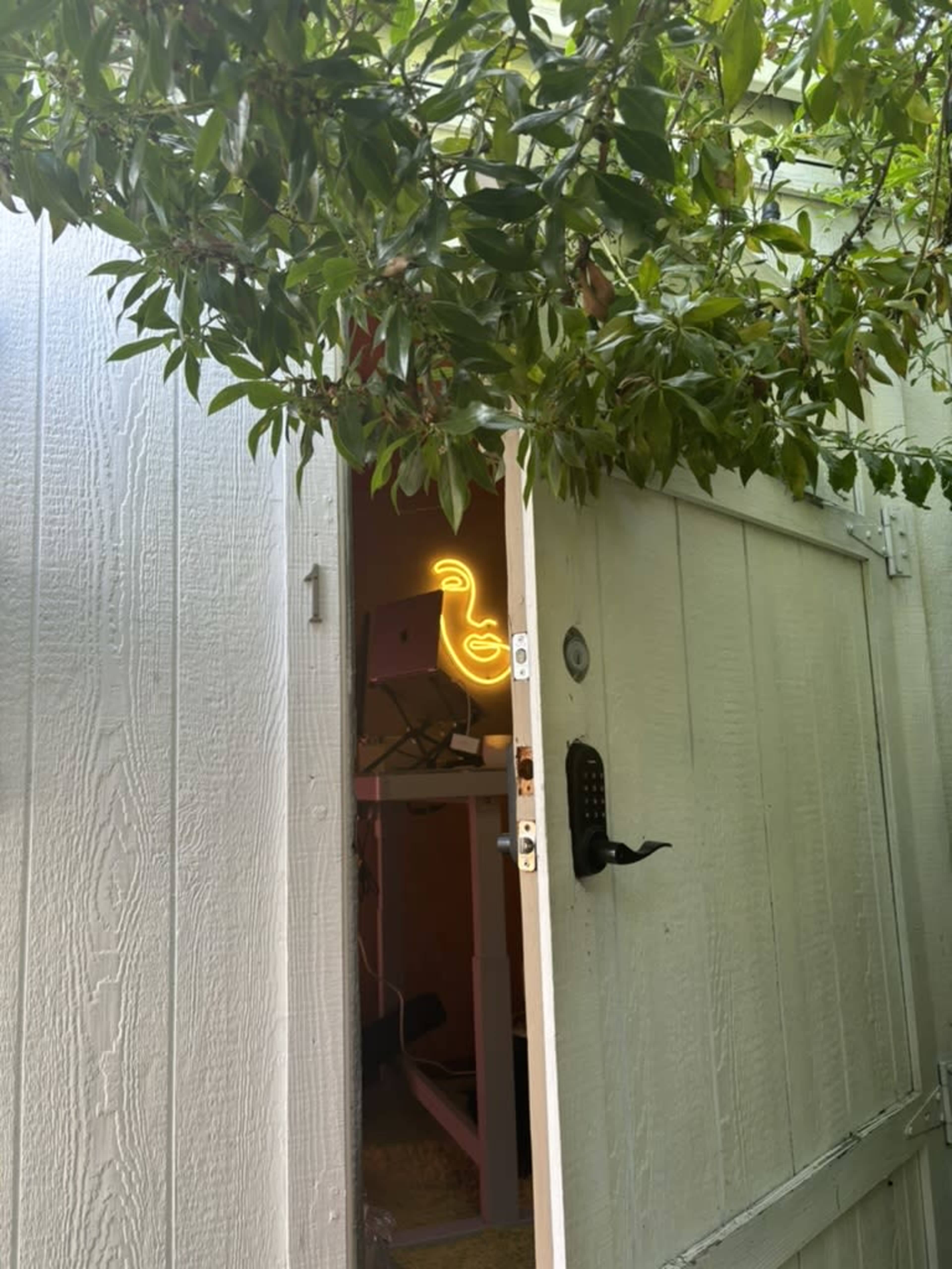 A partially open door reveals a room with a glowing neon duck sign partially obscured by tree foliage.