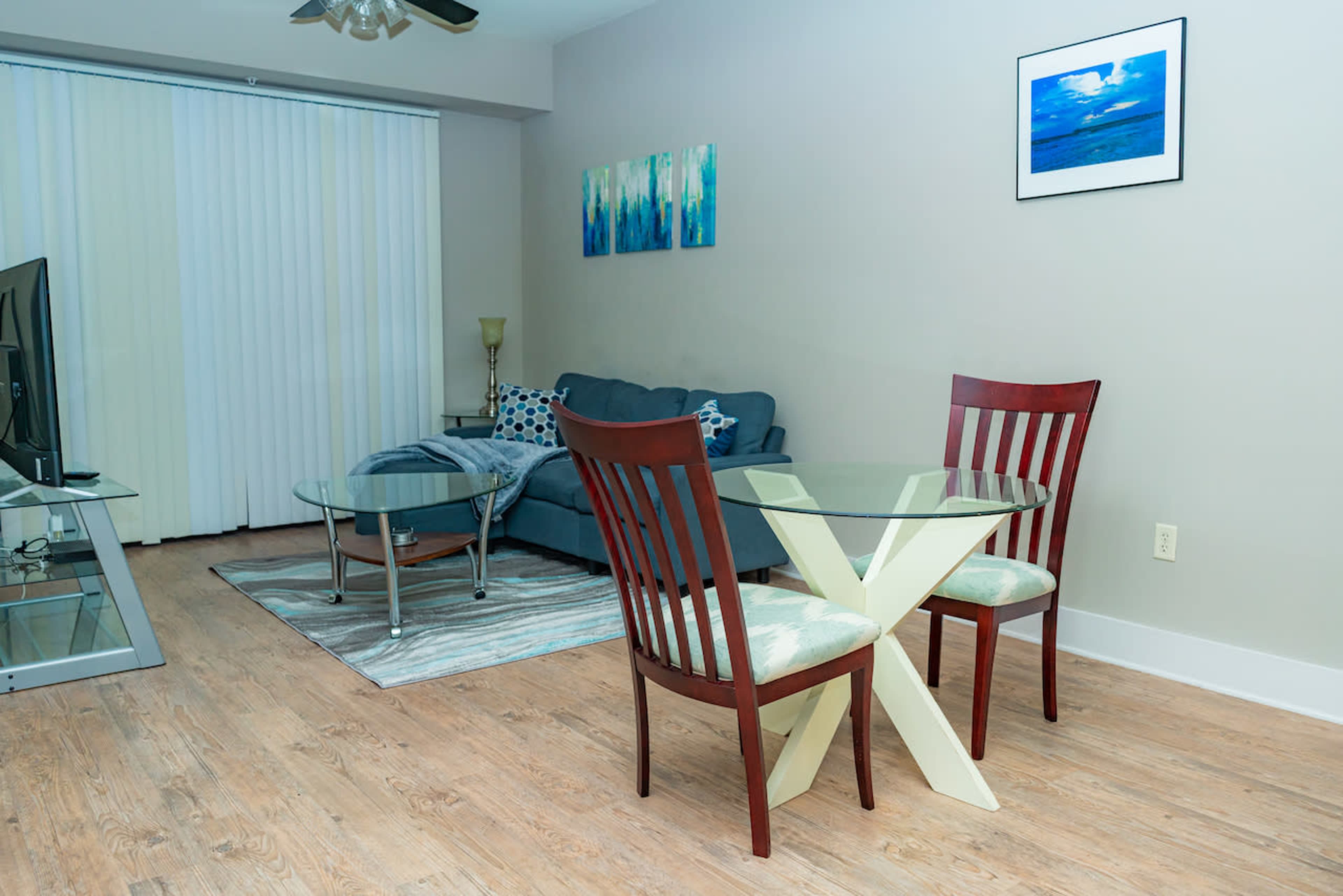 A modern living space features a glass dining table with two chairs, a blue couch, decorative wall art, and large windows with sheer blinds.