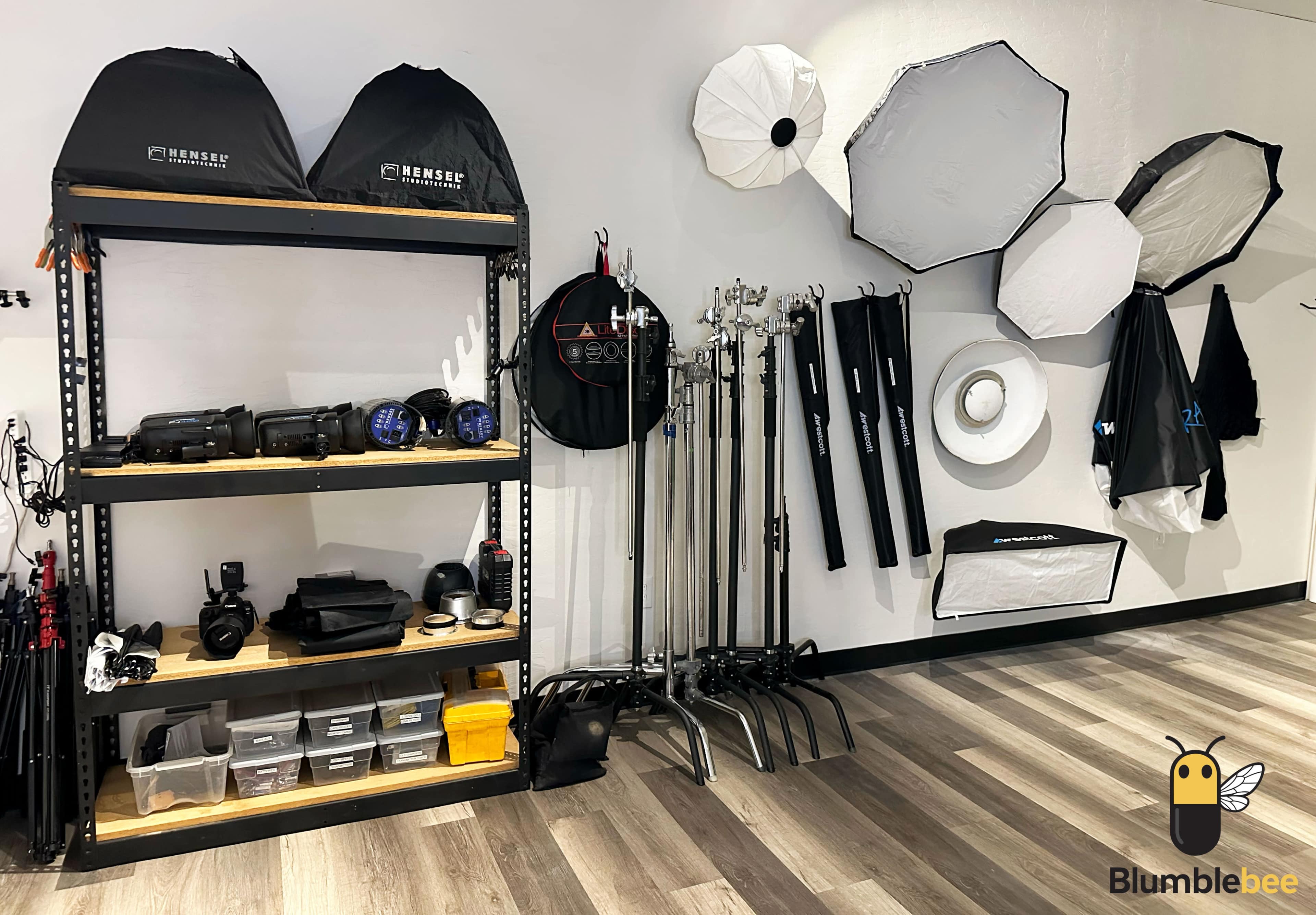 The image shows a neatly organized photography studio with equipment stored on shelves and various light modifiers hanging on the wall.