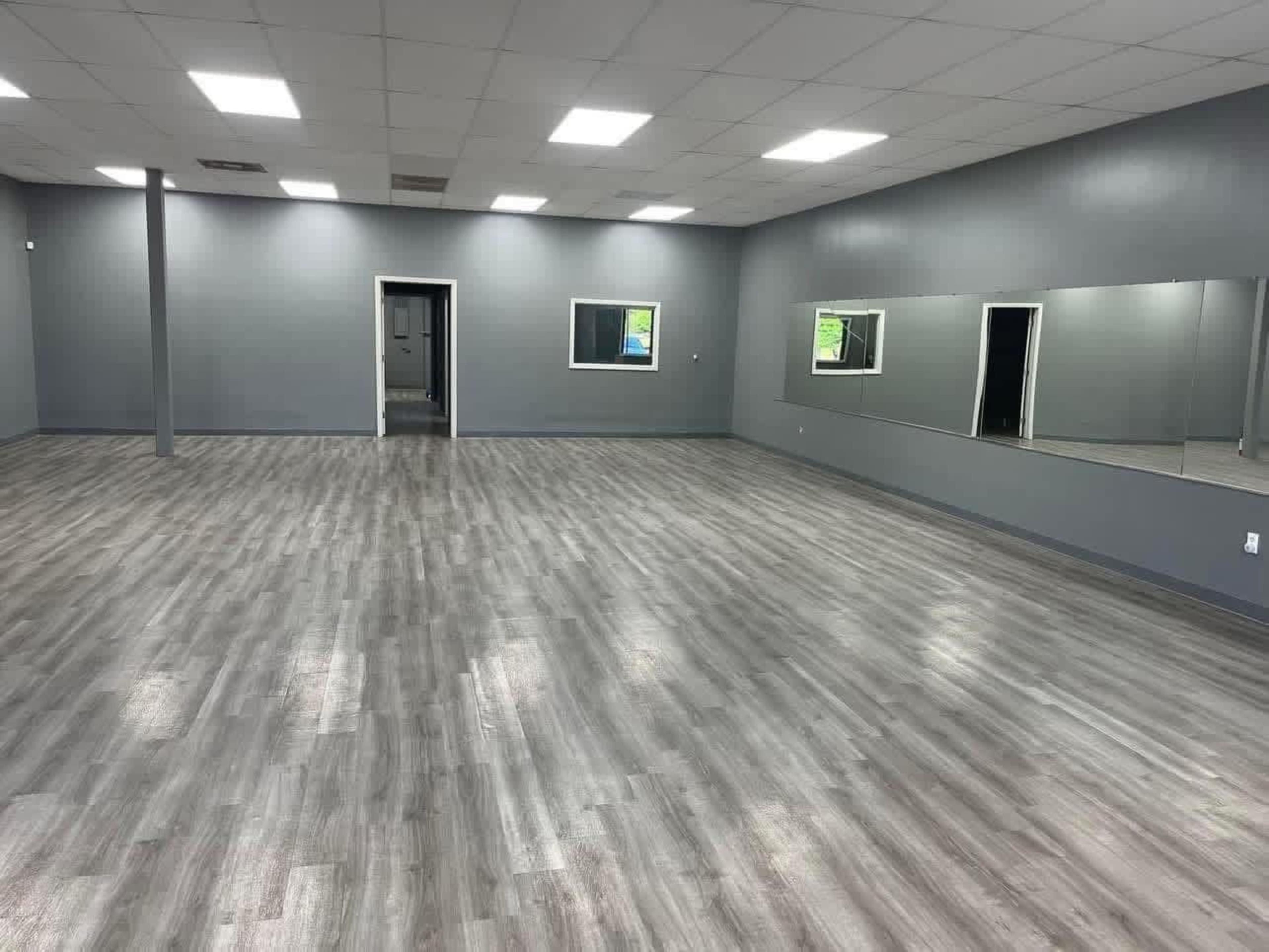 The image shows a spacious, empty room with gray walls, a mirrored wall, and vinyl flooring.