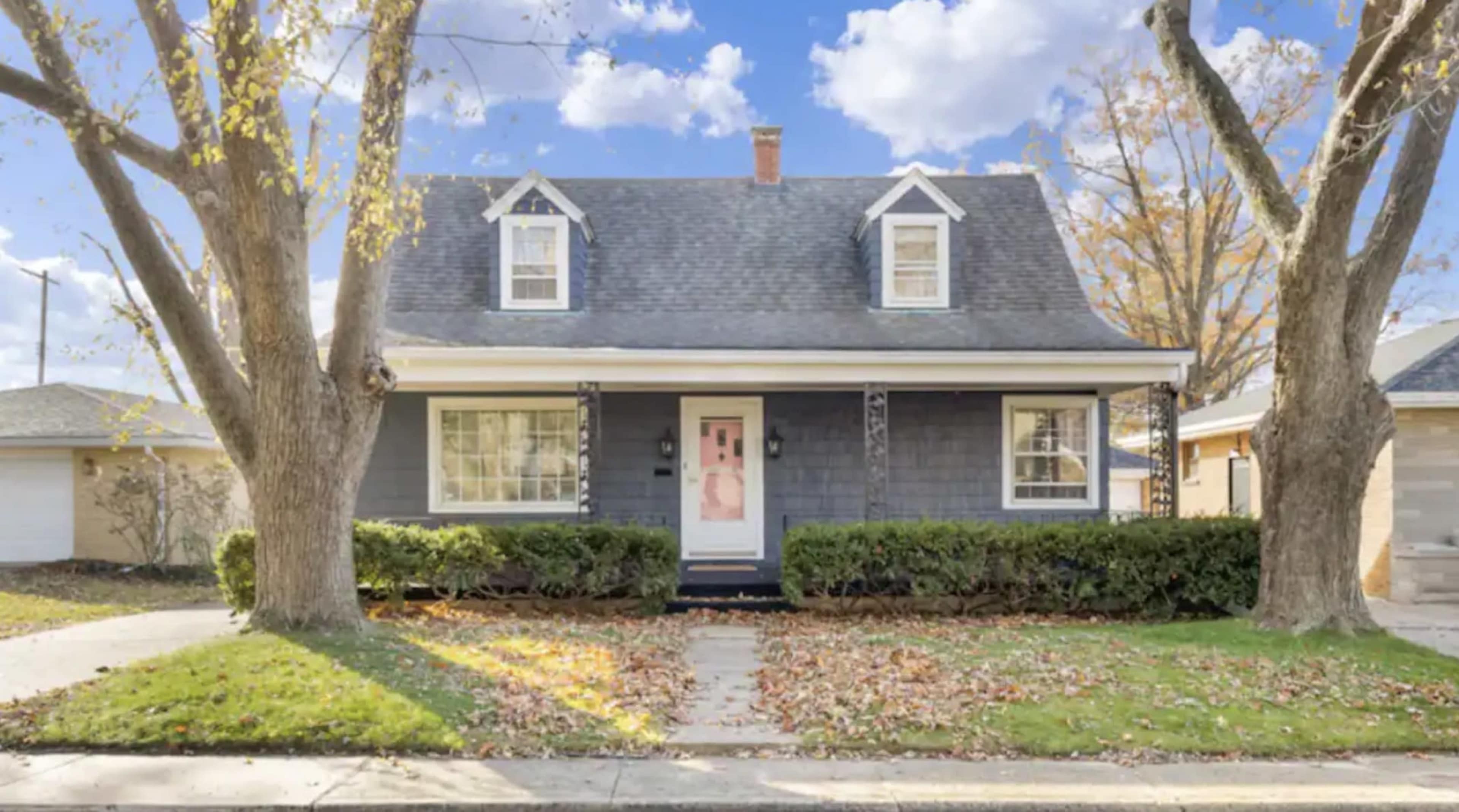 A gray, two-story house with a gabled roof, white trim, and a front porch is surrounded by neatly trimmed bushes and scattered autumn leaves.