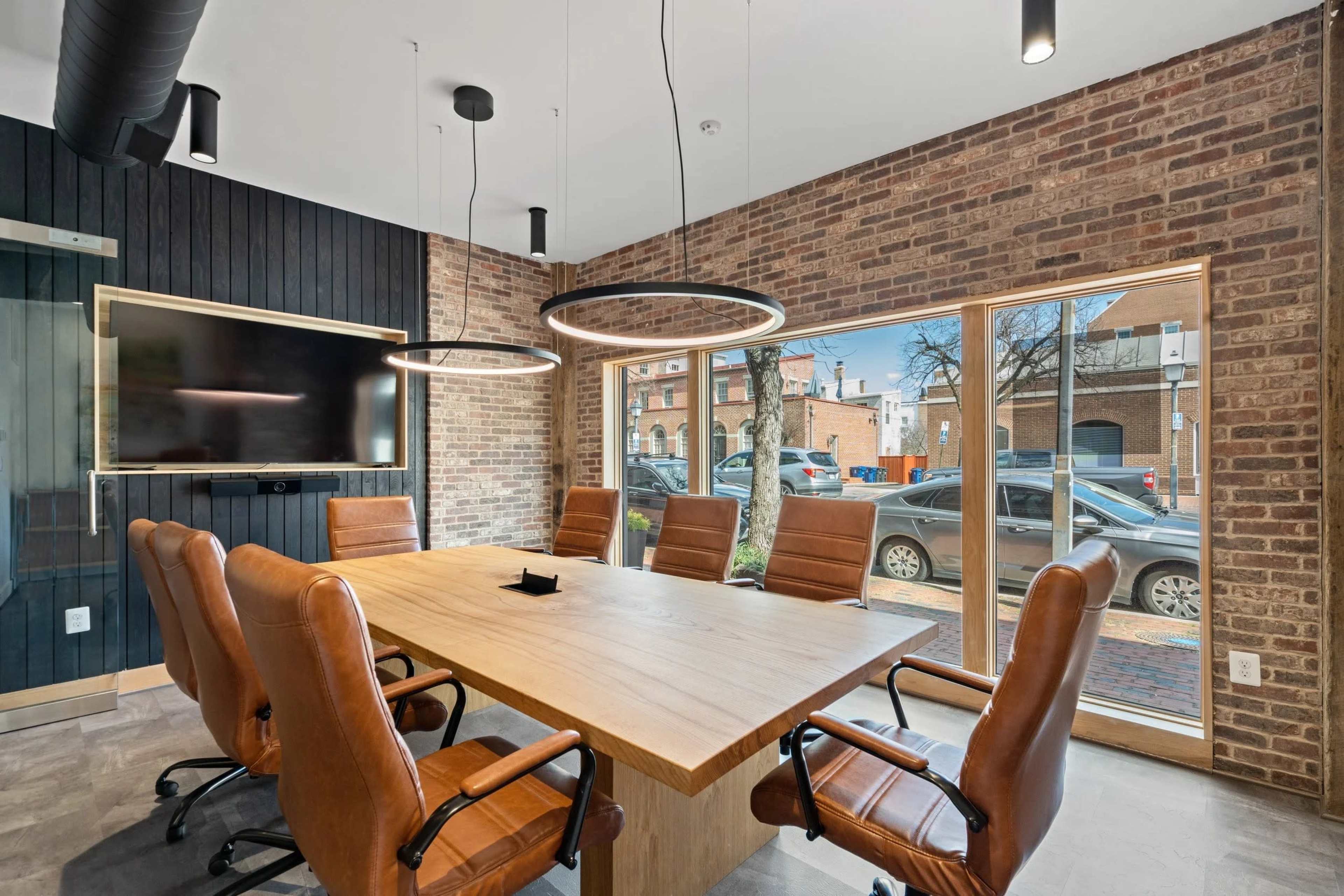 The image shows a modern conference room with a large wooden table surrounded by brown leather chairs, featuring exposed brick walls and large windows overlooking an urban street.