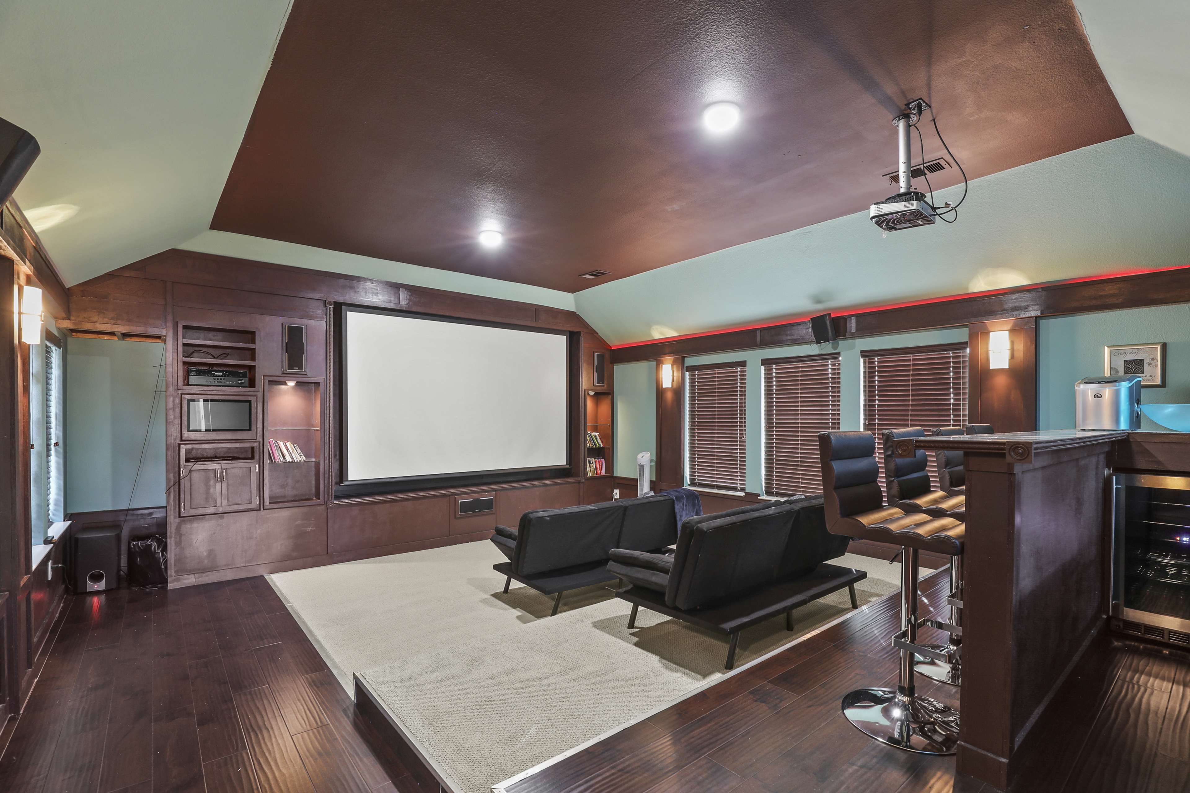 A home theater with a large screen at the front, seating arranged in two black chairs, and a bar area on one side.