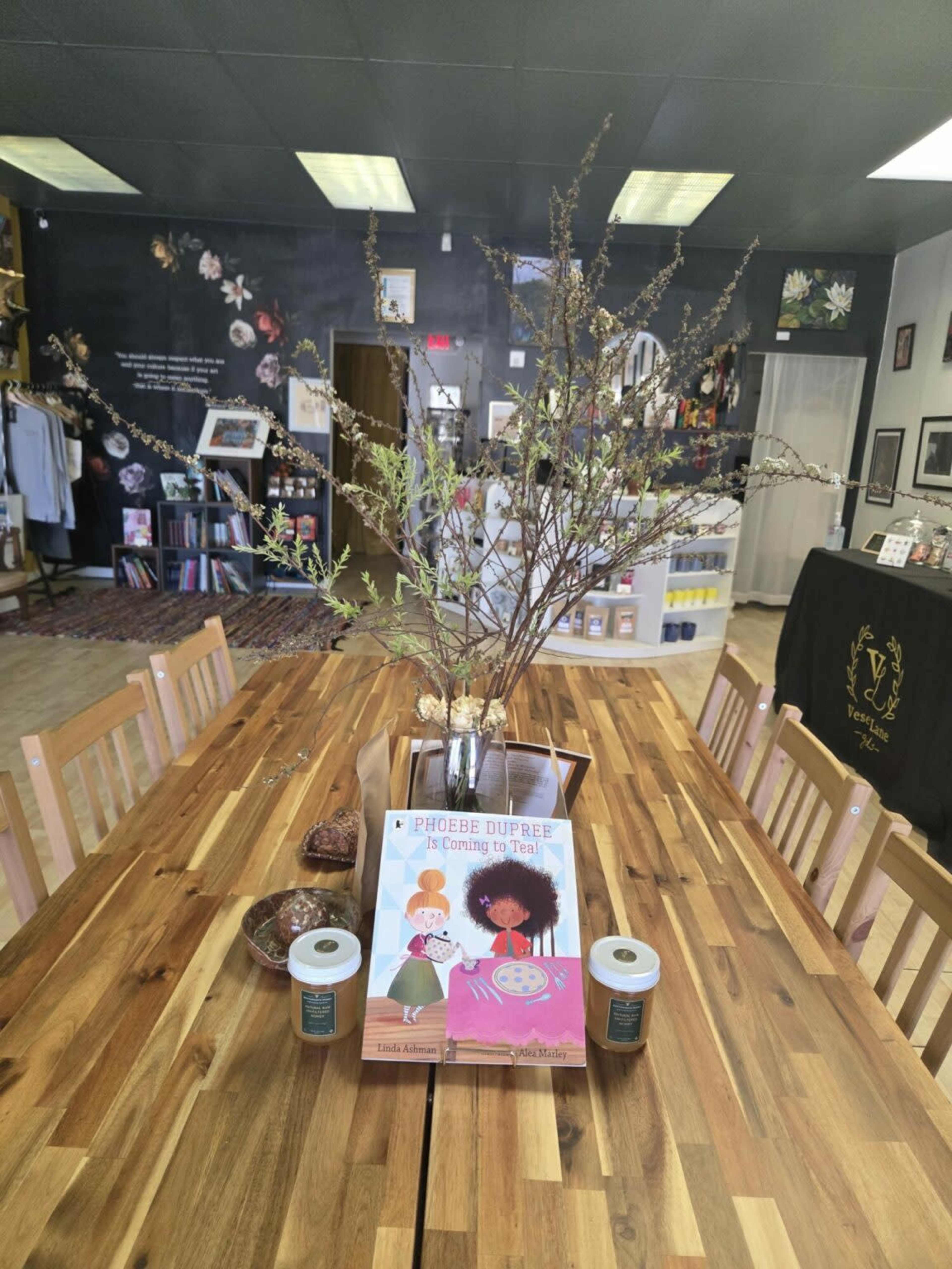 A wooden table is displayed in a shop, featuring a flower arrangement, a book titled "Phoebe Dupree is Coming to Tea," and two candles.