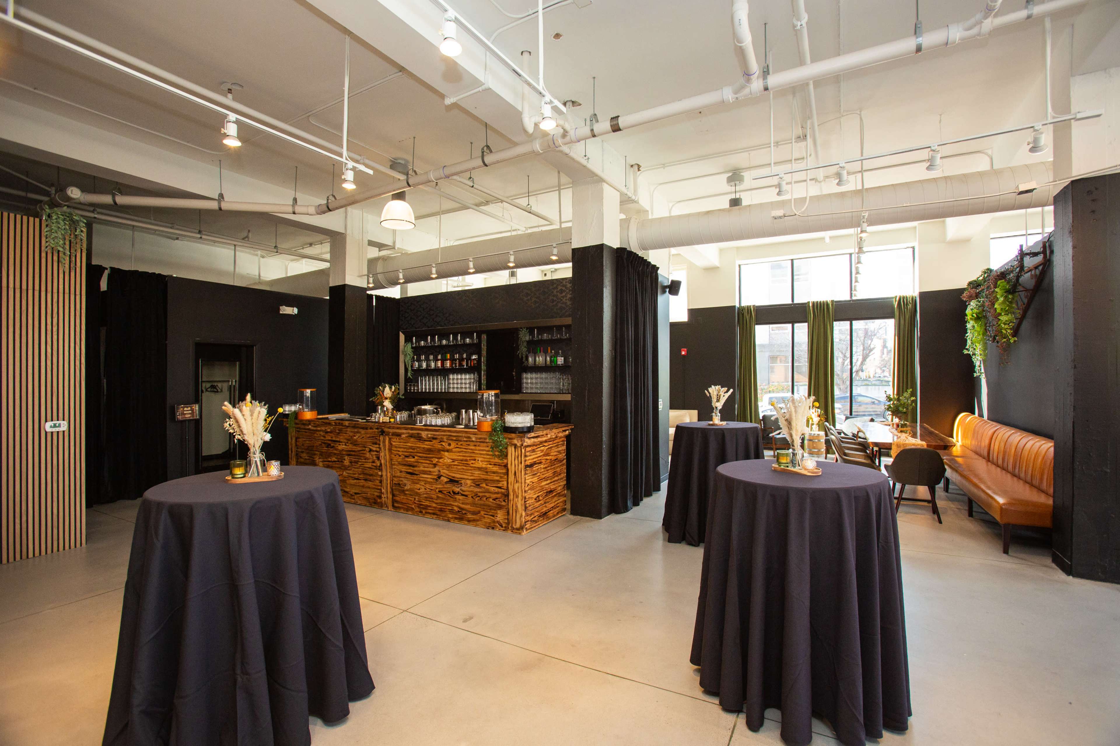 The image shows a modern event space featuring round tables with dark tablecloths, a wooden bar area, and a large window allowing natural light to enter.