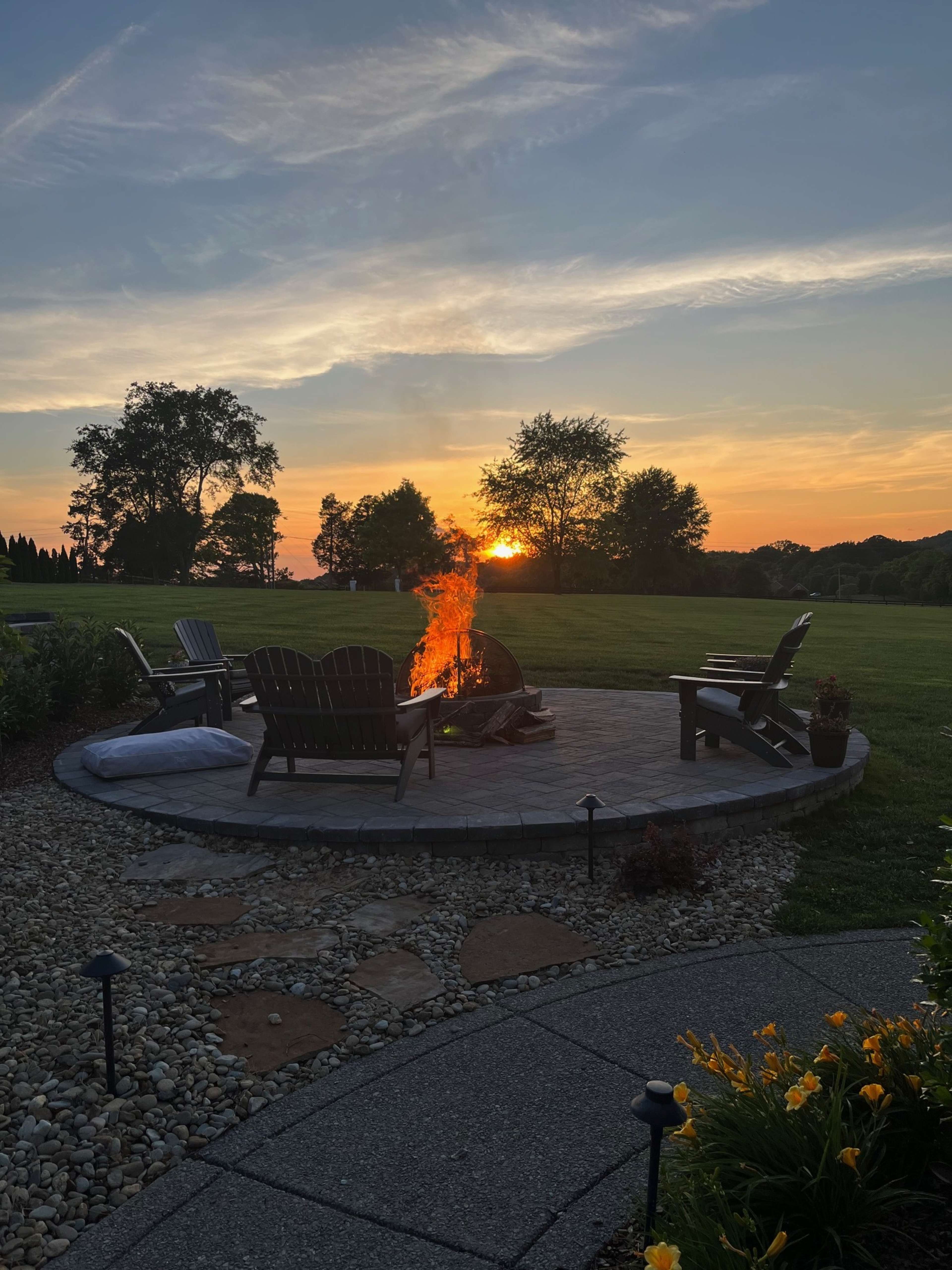 A fire burns in a stone fire pit surrounded by wooden chairs as the sun sets in the background over a grassy field.