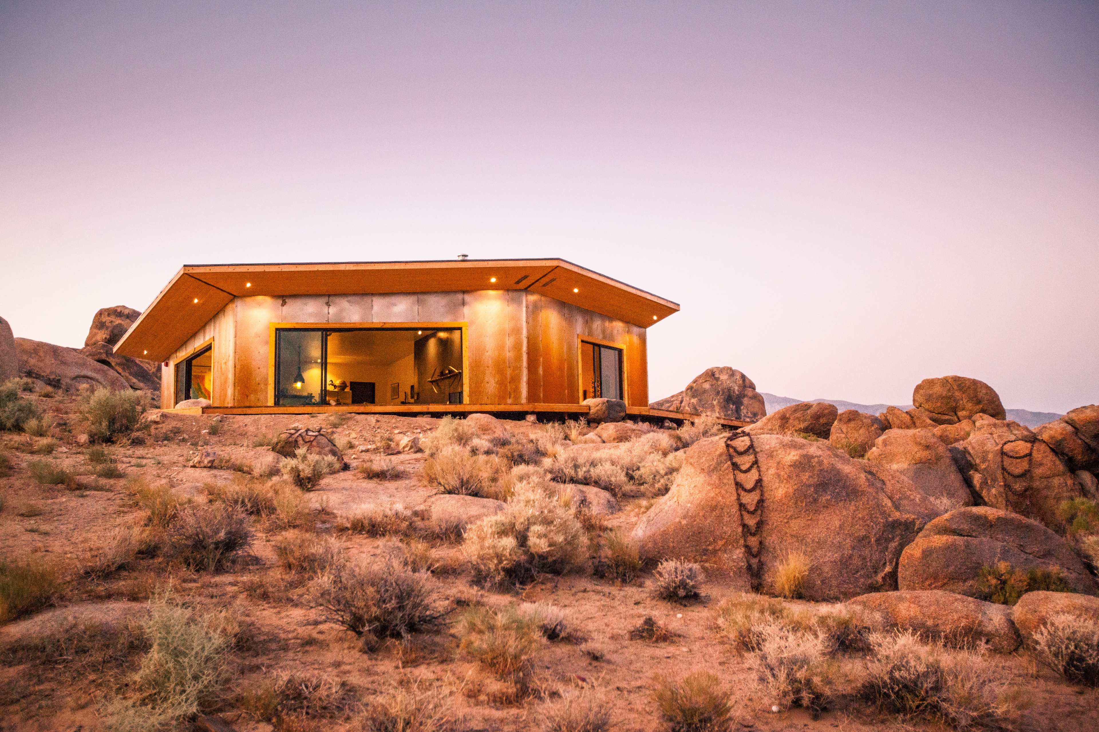 A modern house with large windows sits atop a rocky hill, surrounded by sparse vegetation under a twilight sky.
