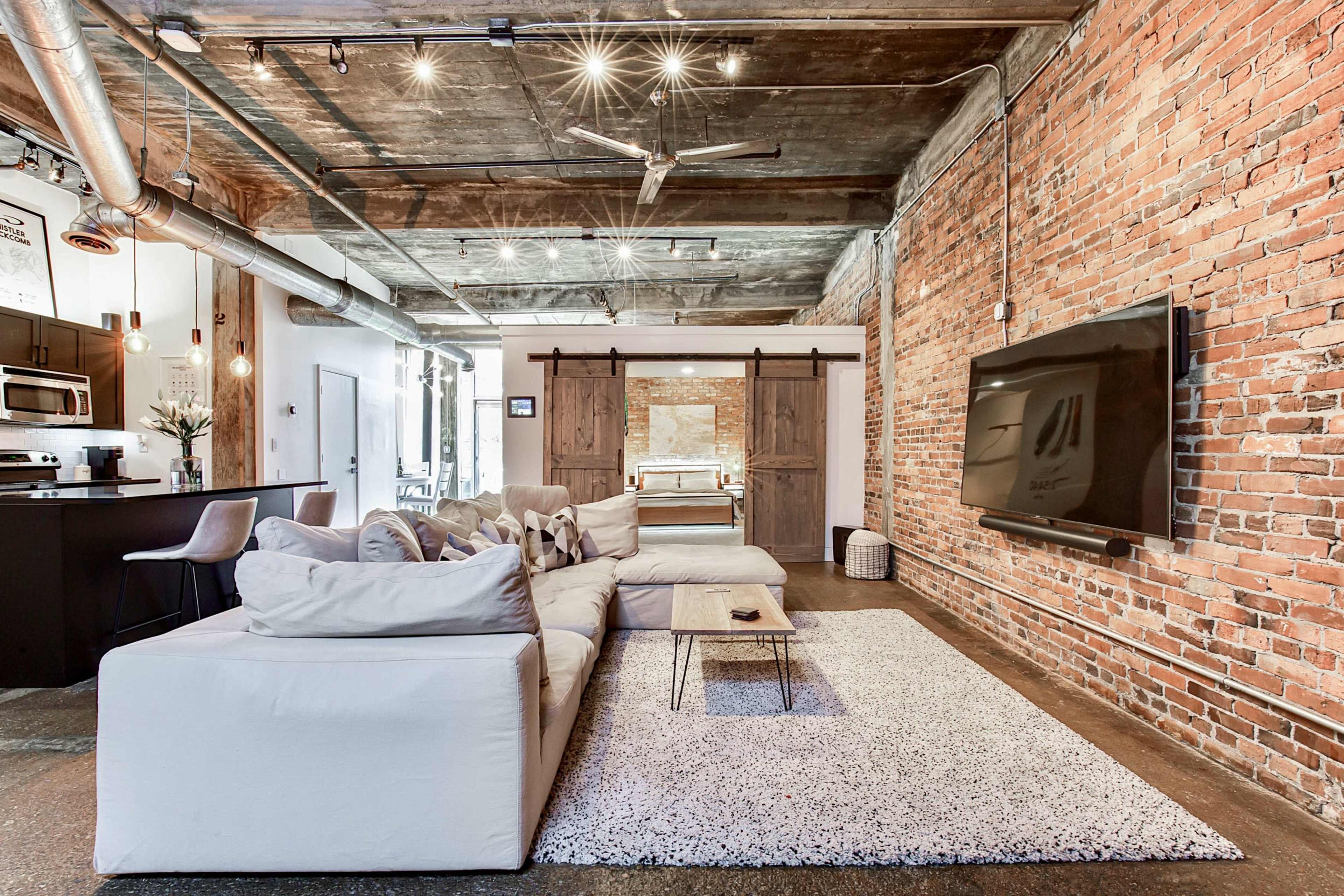 A spacious loft-style living area with exposed brick walls, a large sectional sofa, and a flat-screen TV mounted on the wall, along with a kitchen area visible in the background.