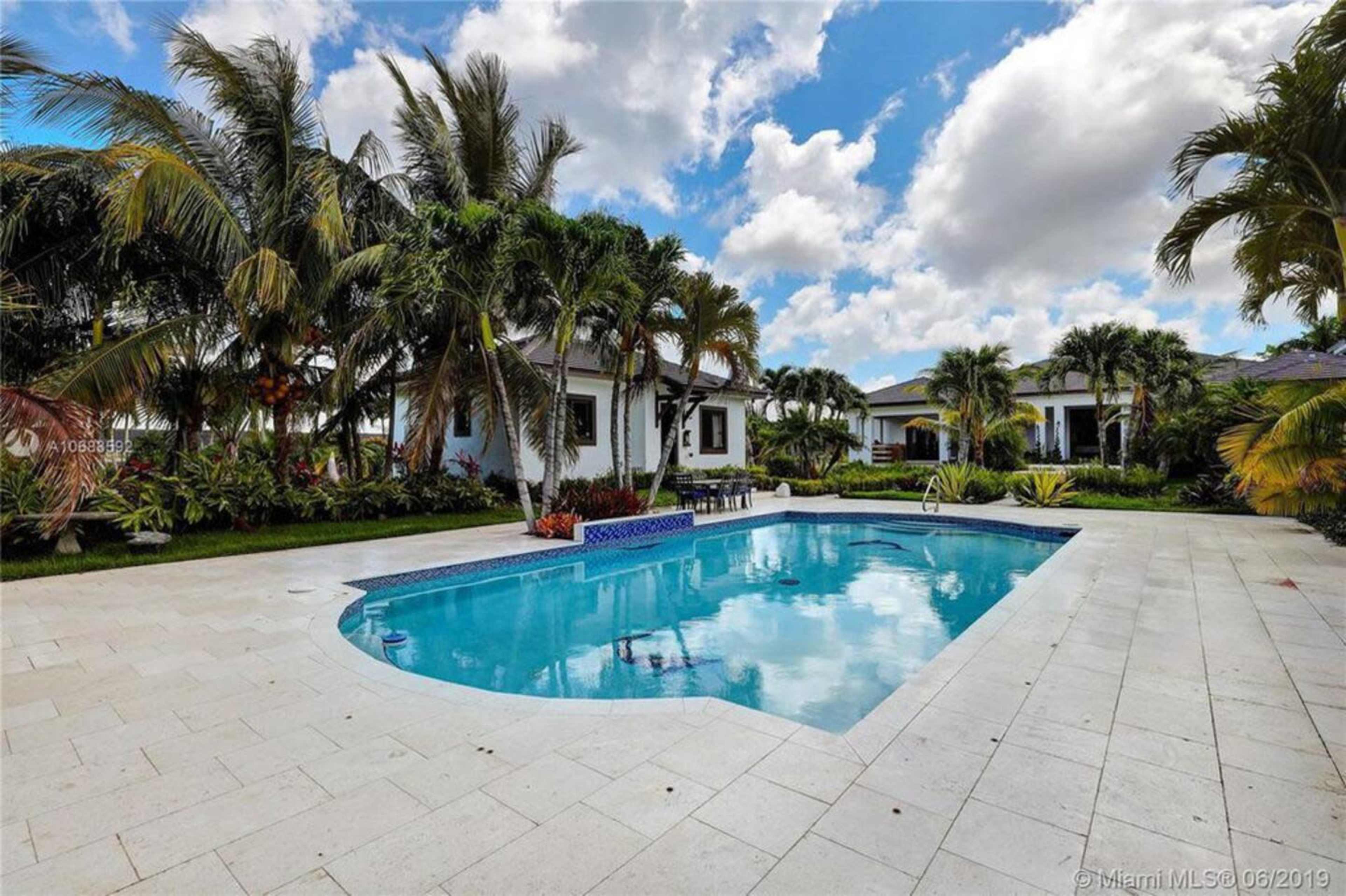 A swimming pool is surrounded by palm trees and landscaped greenery in a residential outdoor area.