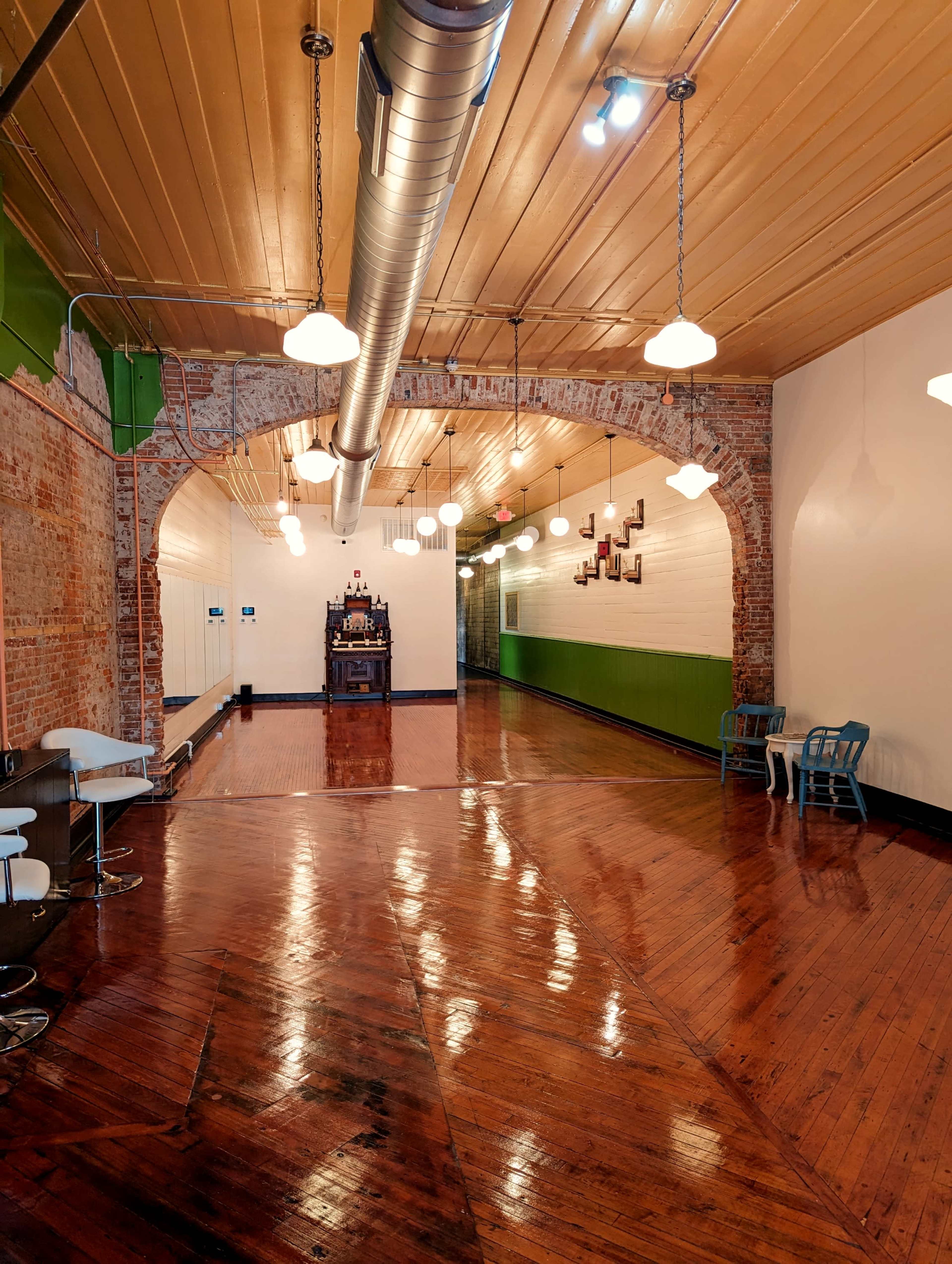 A spacious room with polished wooden floors, exposed brick walls, and a row of pendant lights, featuring a decorative wall at one end.