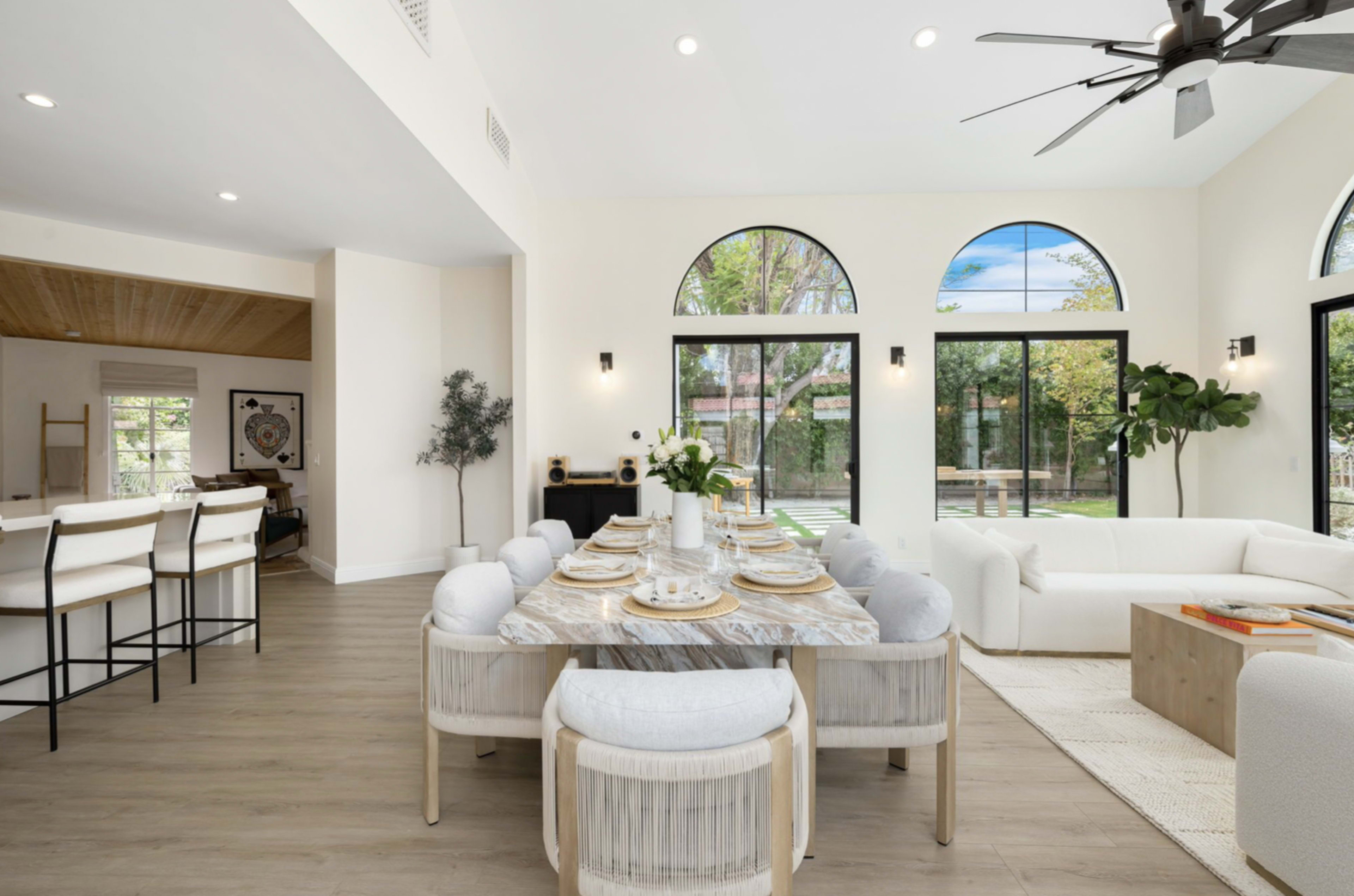 A modern dining area features a round marble table set for six, surrounded by light-colored upholstered chairs, with large windows providing natural light and a view of an outdoor garden.