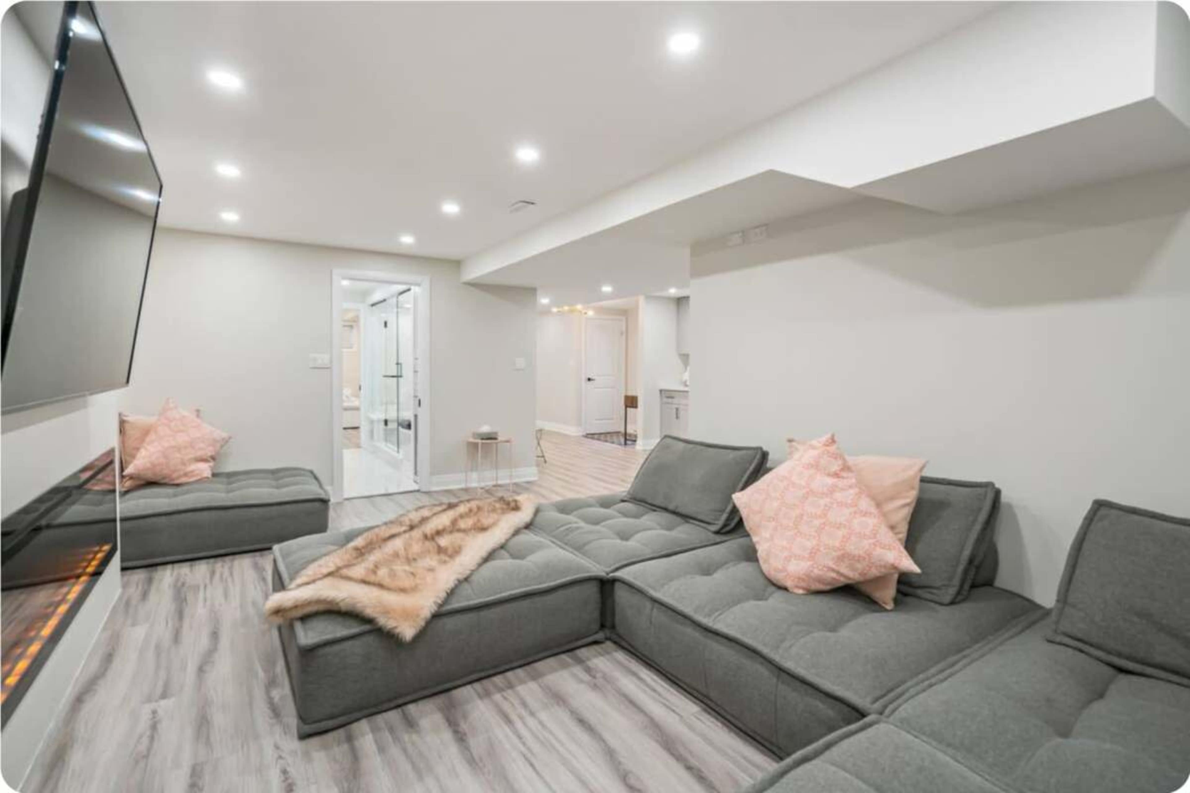 A modern living room features a gray sectional sofa with pink pillows and a wall-mounted TV, adjacent to a glass-enclosed bathroom and kitchen area.