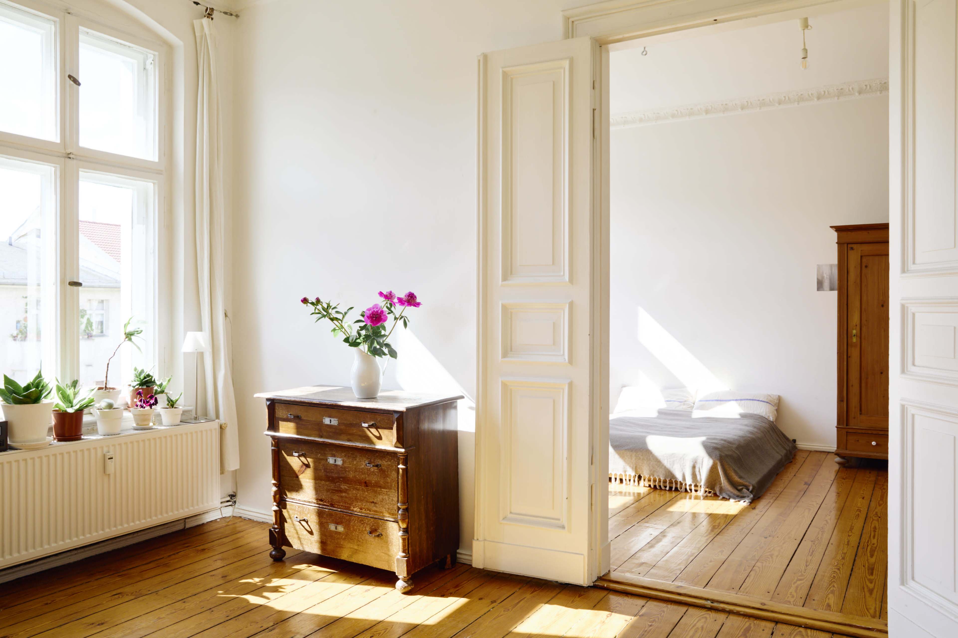 The image shows a bright, airy room with a wooden dresser and potted plants, leading to a second room featuring a bed covered with a grey blanket.
