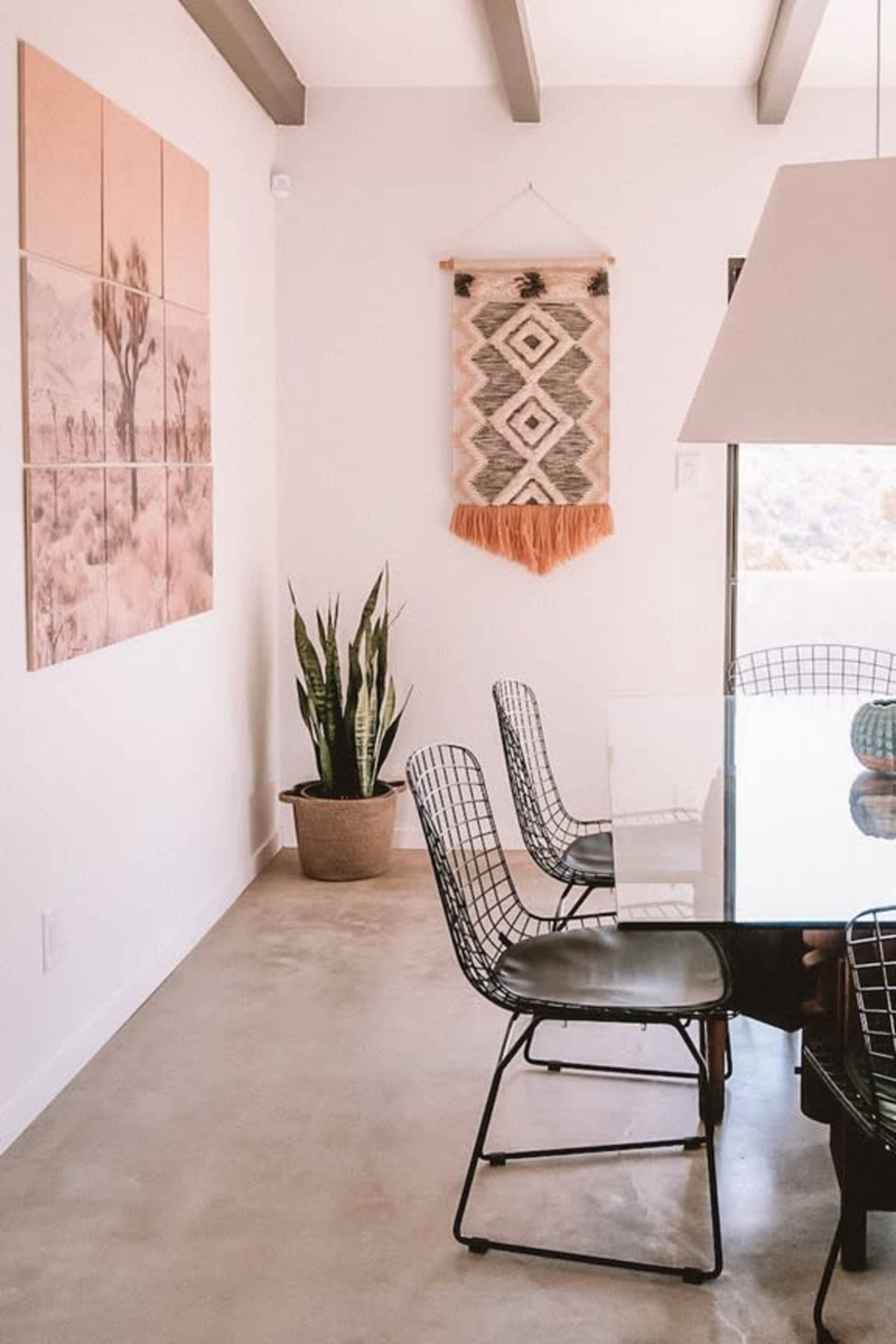 A modern dining area featuring a glass table, black wire chairs, a potted plant, and wall art consisting of photographs and a textile hanging.