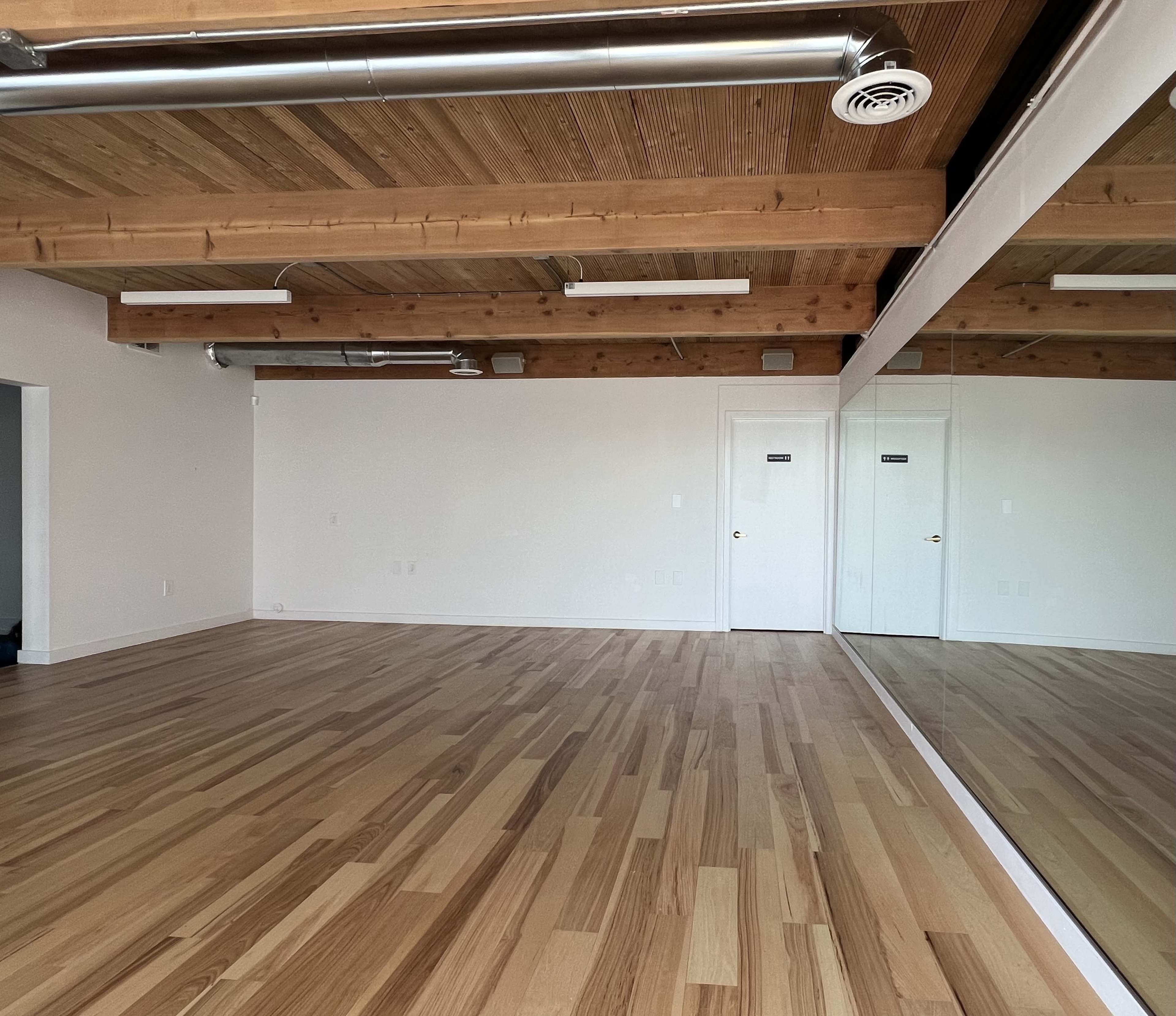 An empty room with hardwood floors and a large mirror on one wall, featuring exposed wooden beams on the ceiling.