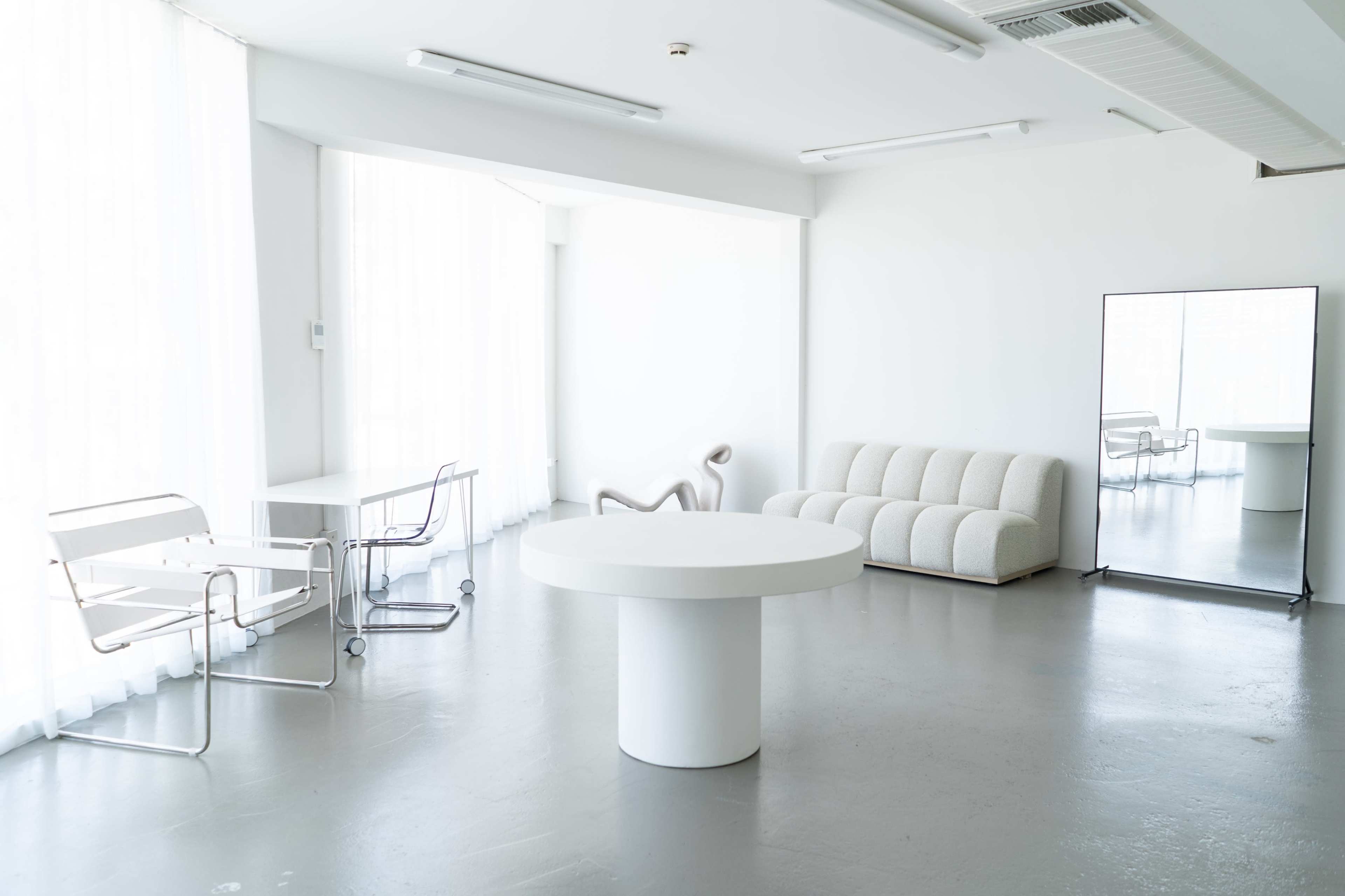 The image shows a modern, minimalist interior with a round white table, a textured sofa, a mirror, and two chairs near large windows with sheer curtains.