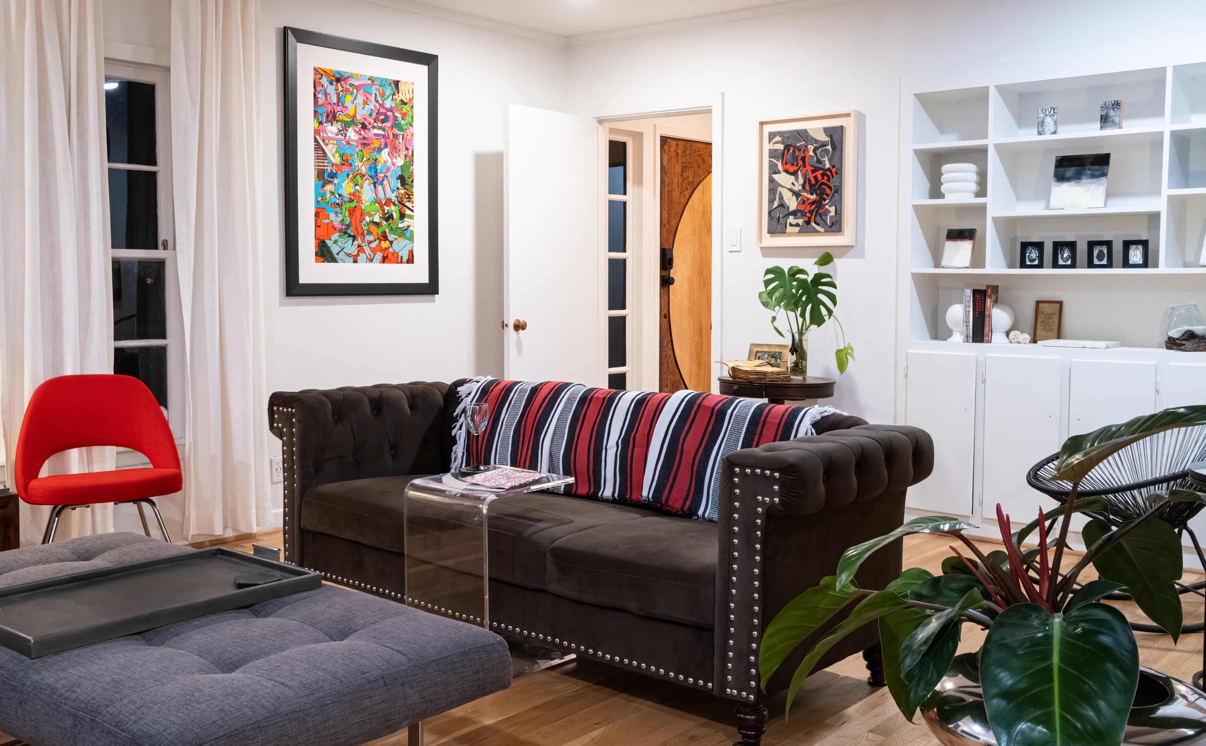 The image shows a living room with a tufted brown sofa adorned with striped cushions, a red chair, a glass coffee table, and various artworks on the walls.