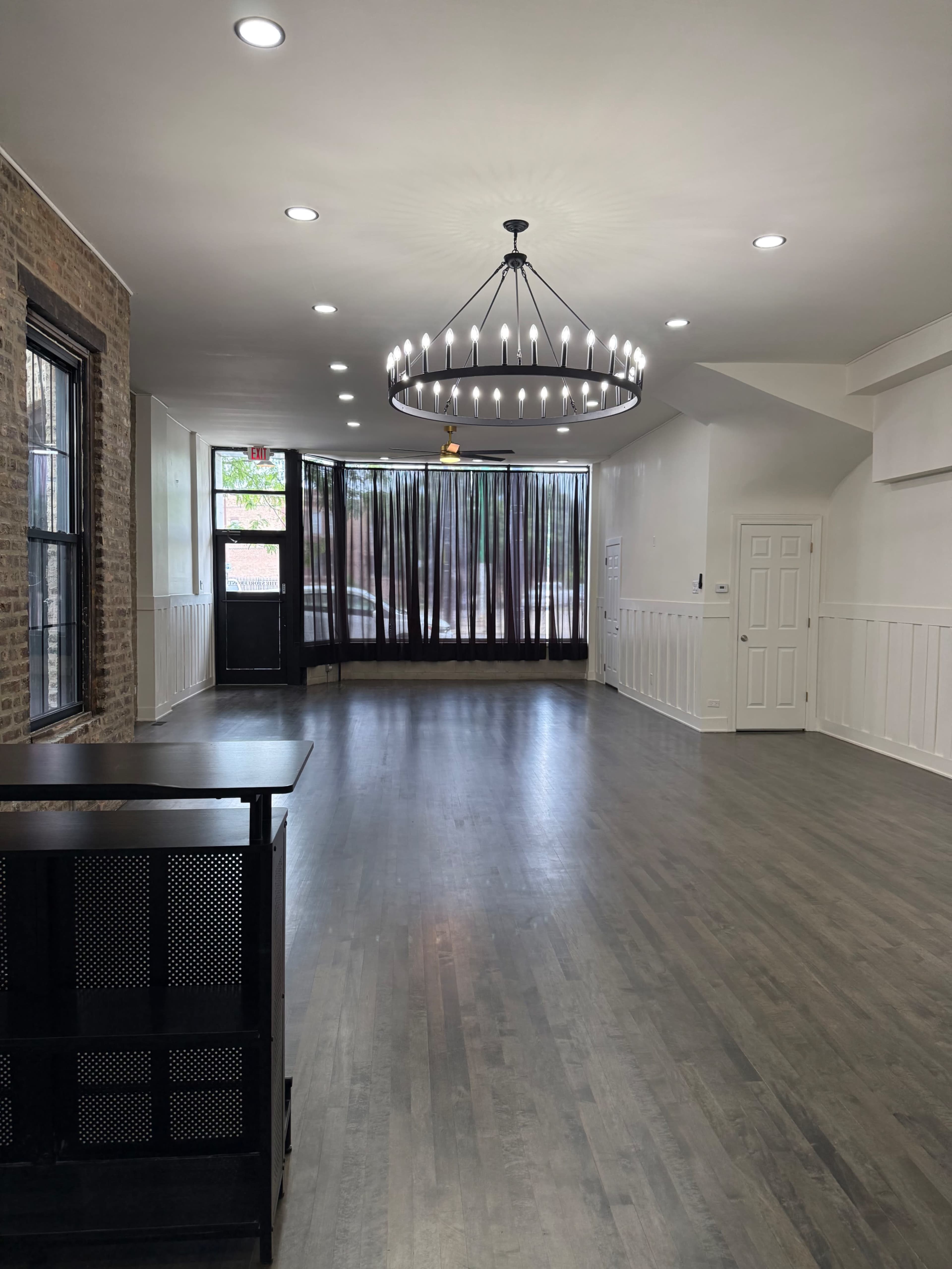 The image shows a spacious interior room with hardwood flooring, large windows covered by dark curtains, and a chandelier hanging from the ceiling.