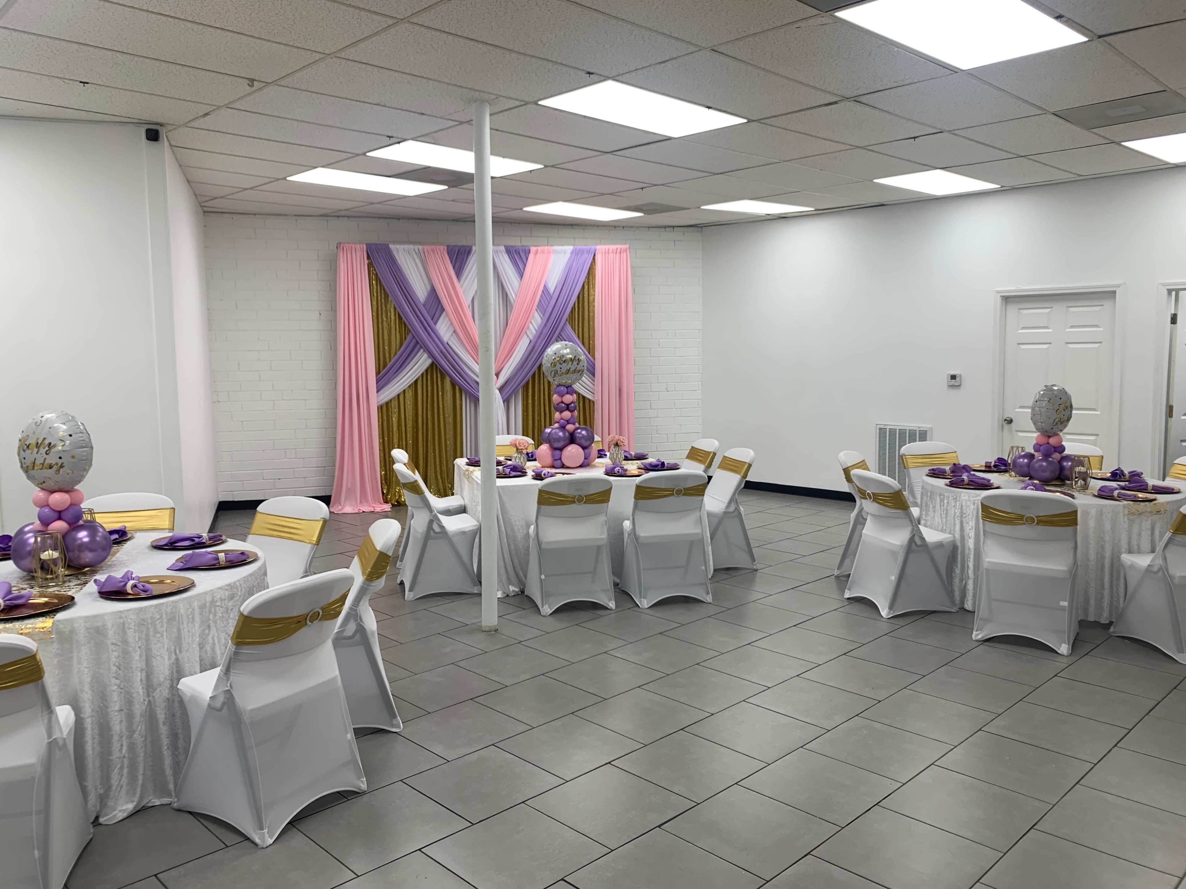 A decorated banquet room with round tables set for an event, featuring purple and gold accents, along with a backdrop of draped fabric.