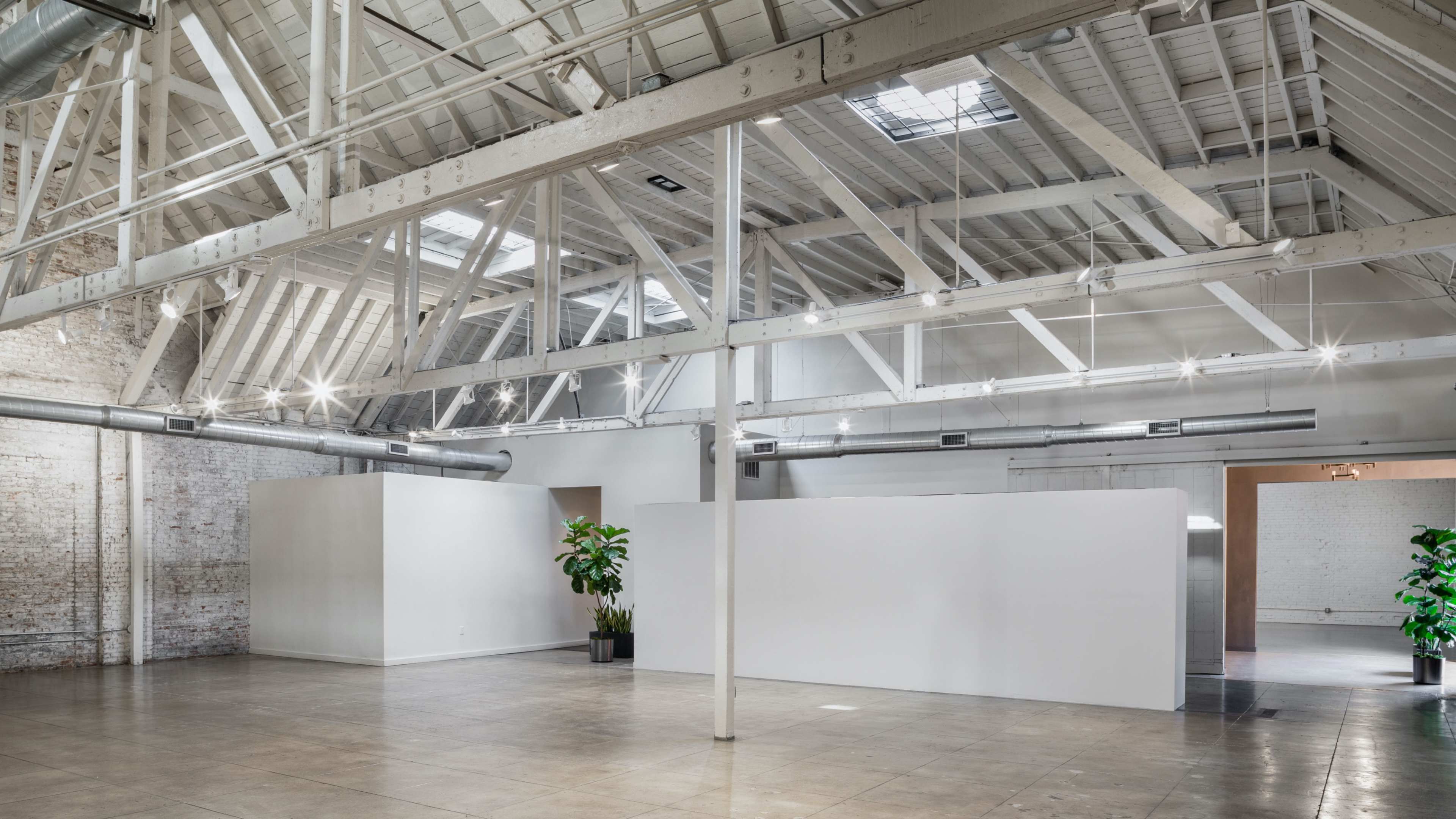 The image shows a spacious, industrial-style interior with high ceilings, exposed beams, and large skylights, featuring white walls and a few potted plants.