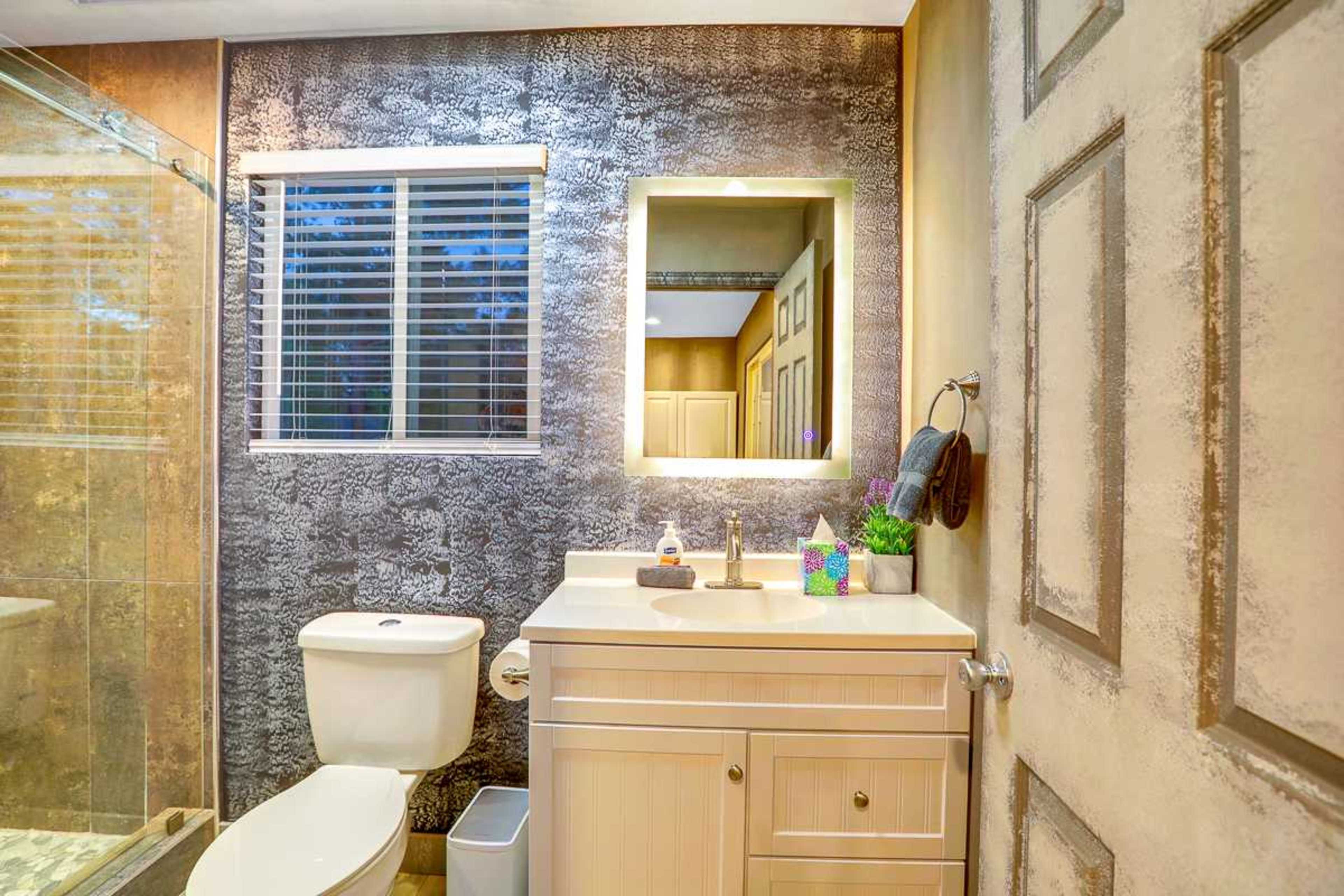A bathroom featuring a toilet, a sink with a countertop, a mirror framed with light, and a glass shower enclosure.