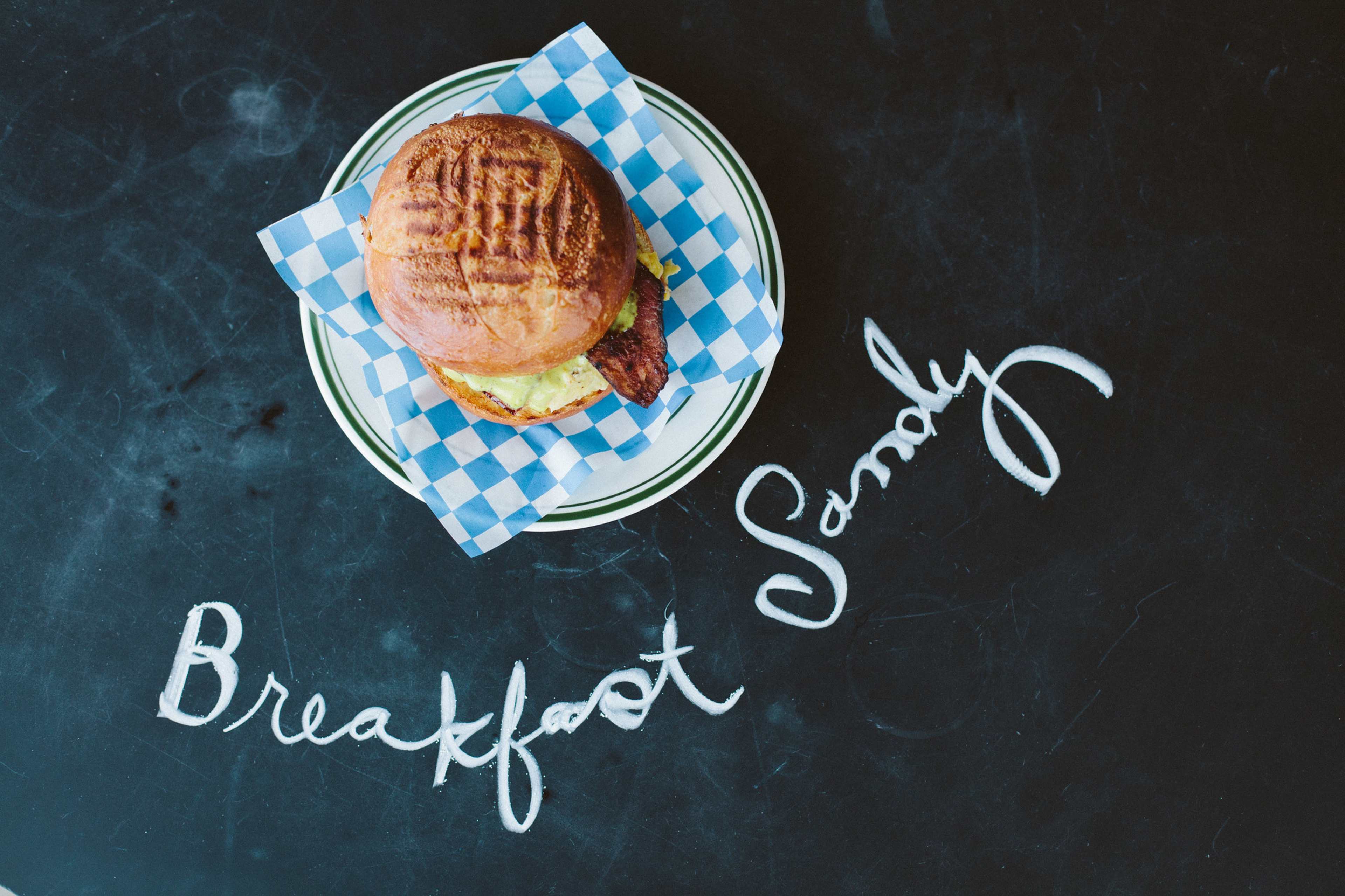 A breakfast sandwich sits on a plate with blue and white checkered paper beside the words "Breakfast Sandy" written in white chalk on a black surface.