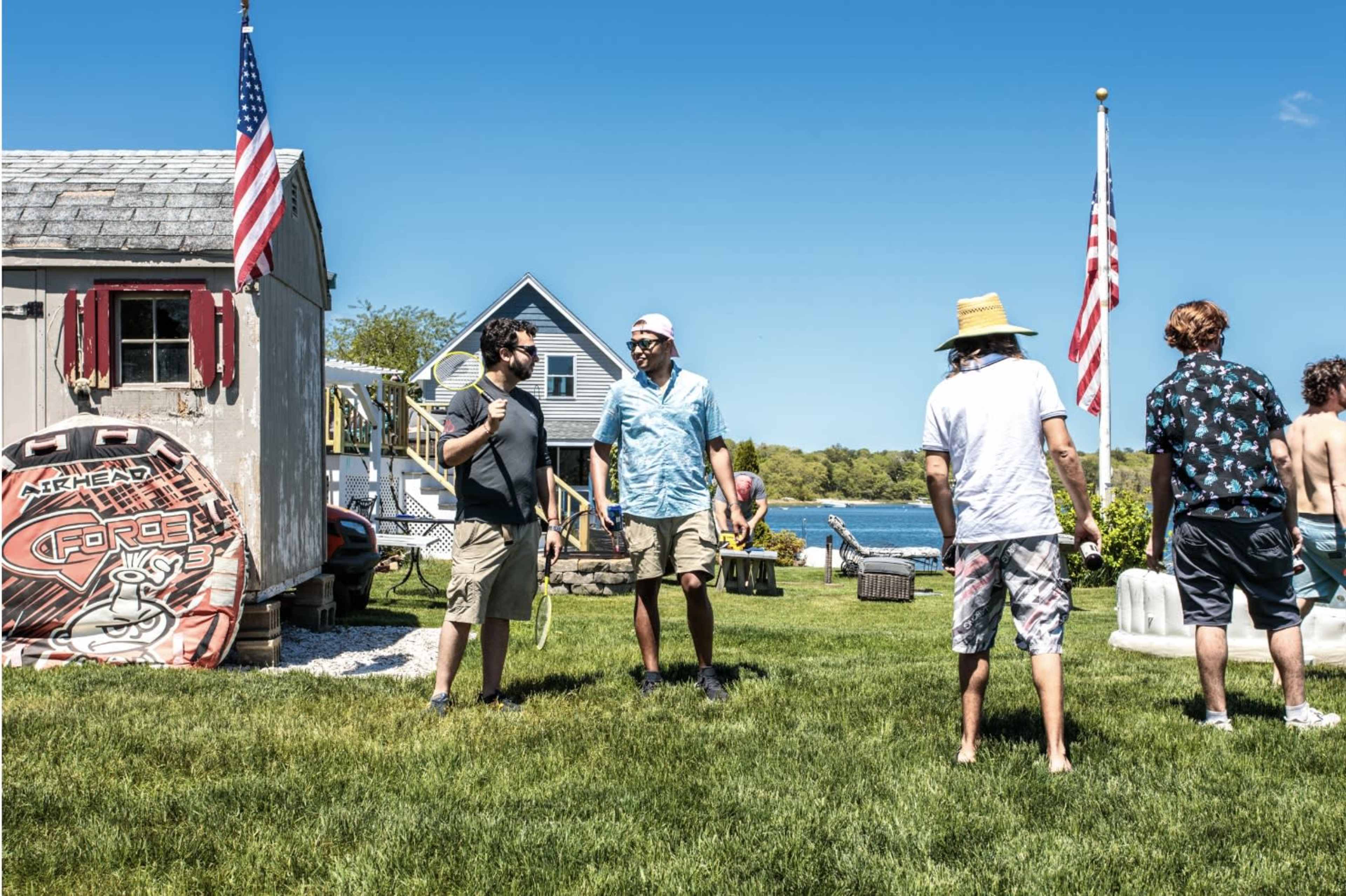 A group of five men stands on a grassy area near a waterfront, with flags and a shed in the background.