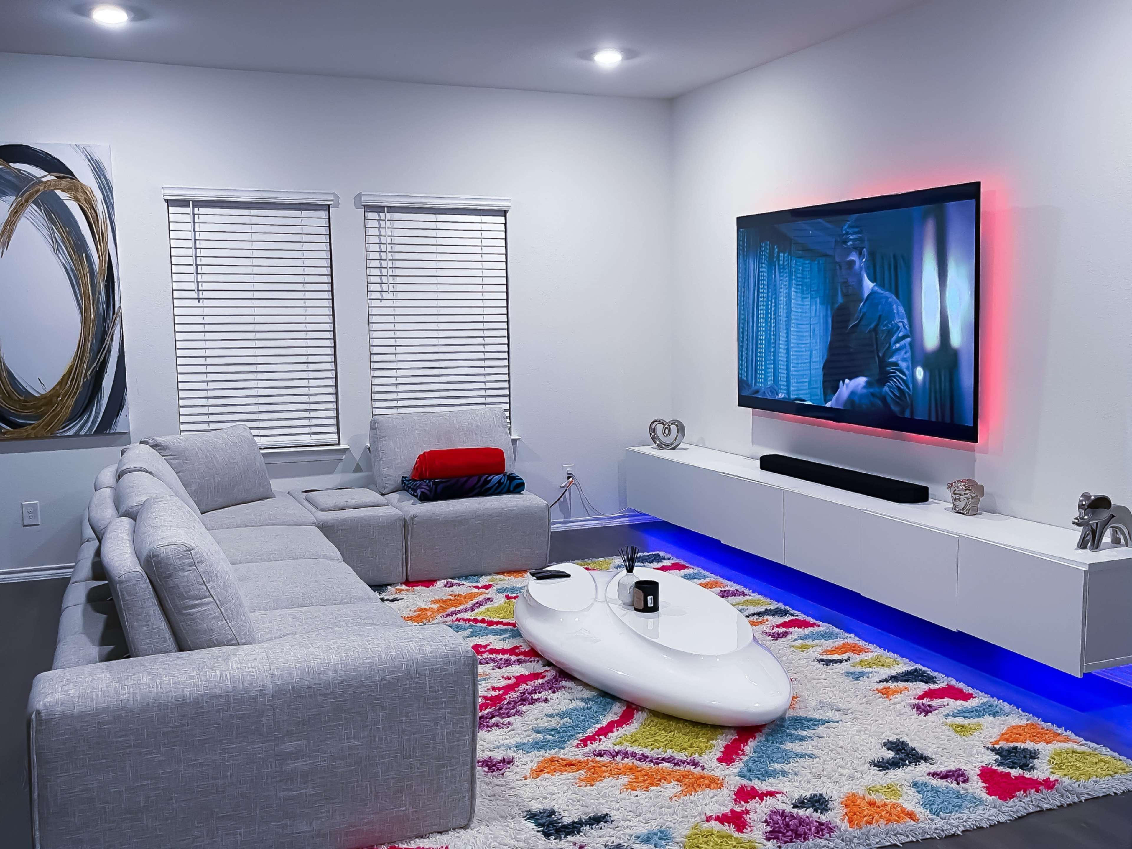 A modern living room features a light gray sectional sofa, a colorful rug with geometric patterns, and a large wall-mounted TV with ambient lighting.