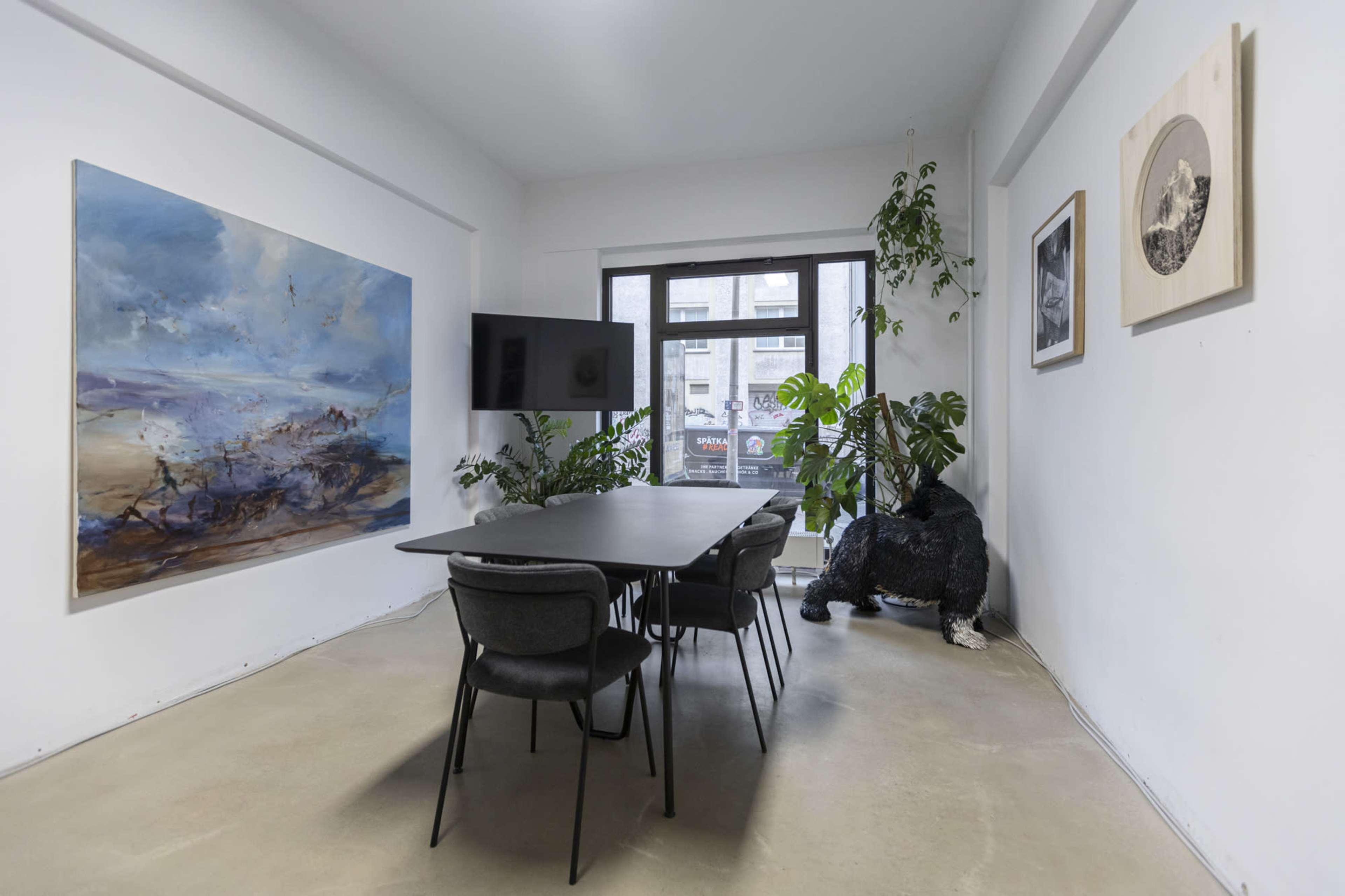 The image shows a modern meeting room with a large painting on the wall, a television, a black table surrounded by chairs, and decorative plants.