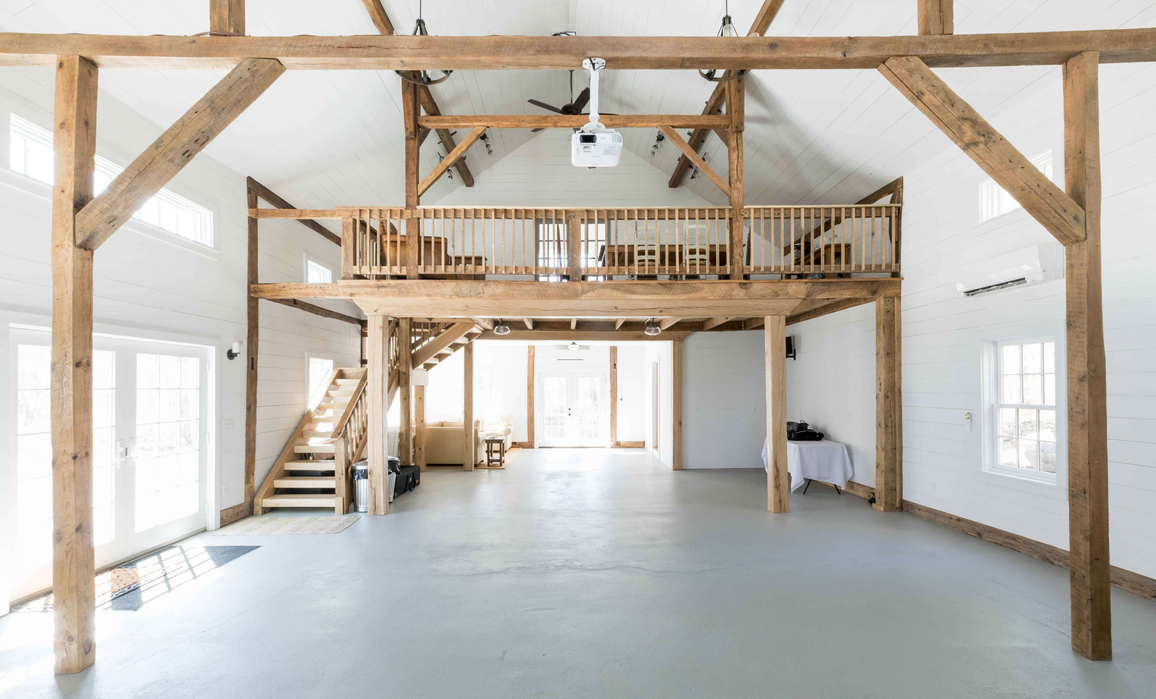 An open interior space featuring exposed wooden beams, a loft area, and large windows that allow natural light to fill the room.