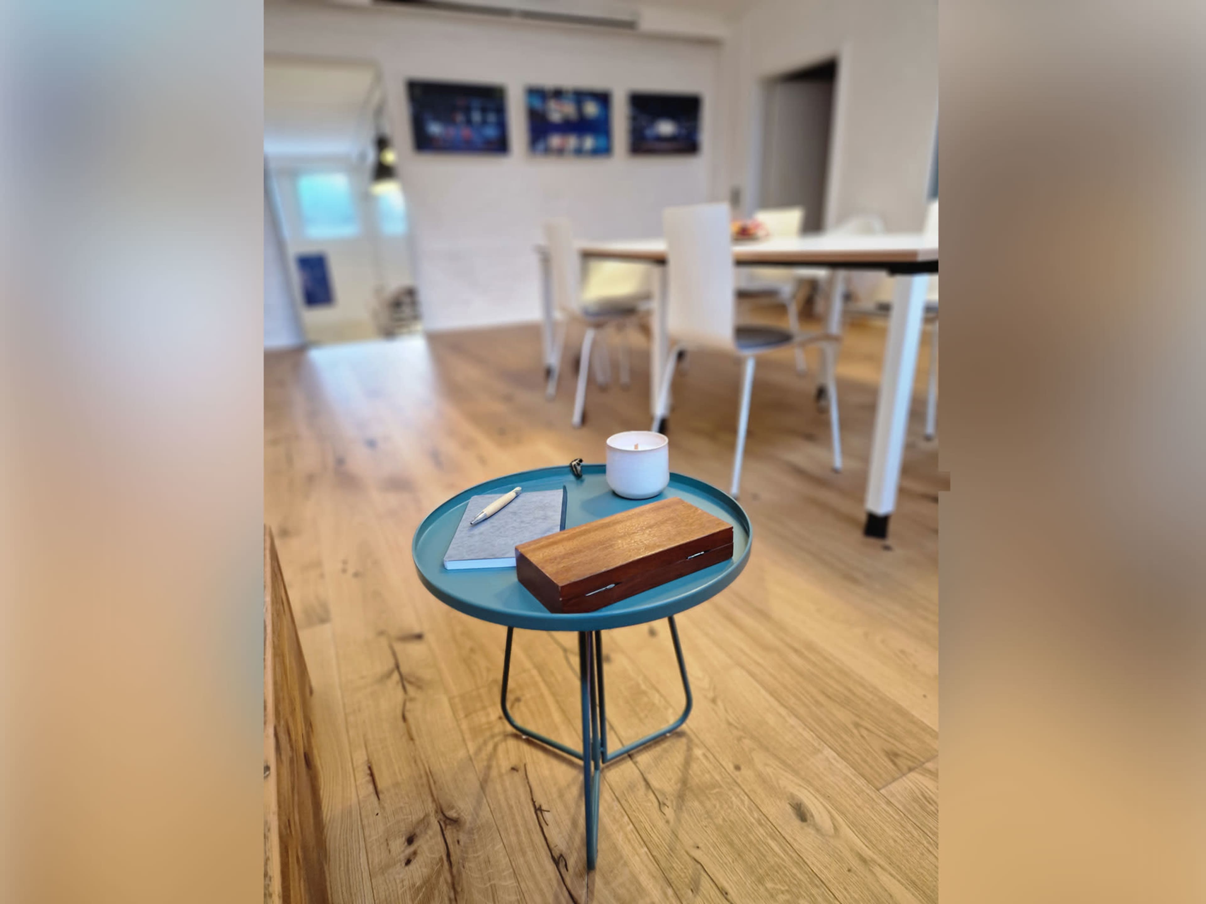 A small round table with a teal top holds a notebook, a pen, and a candle, while a wooden box sits beside them in a well-lit room with white walls and a dining area in the background.