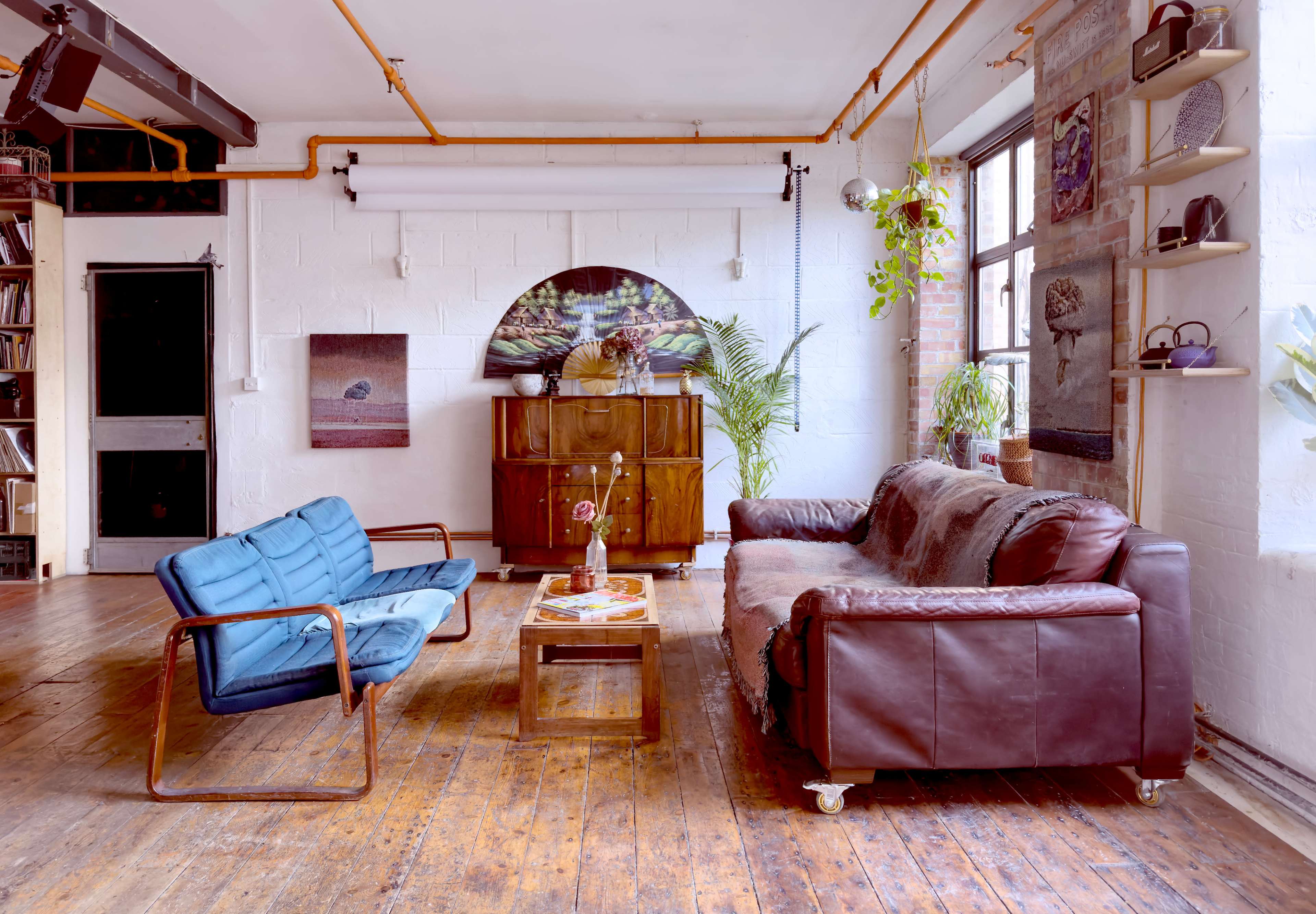 A living room features a blue armchair and a brown leather sofa set against a backdrop of wooden flooring and a large window with plants.