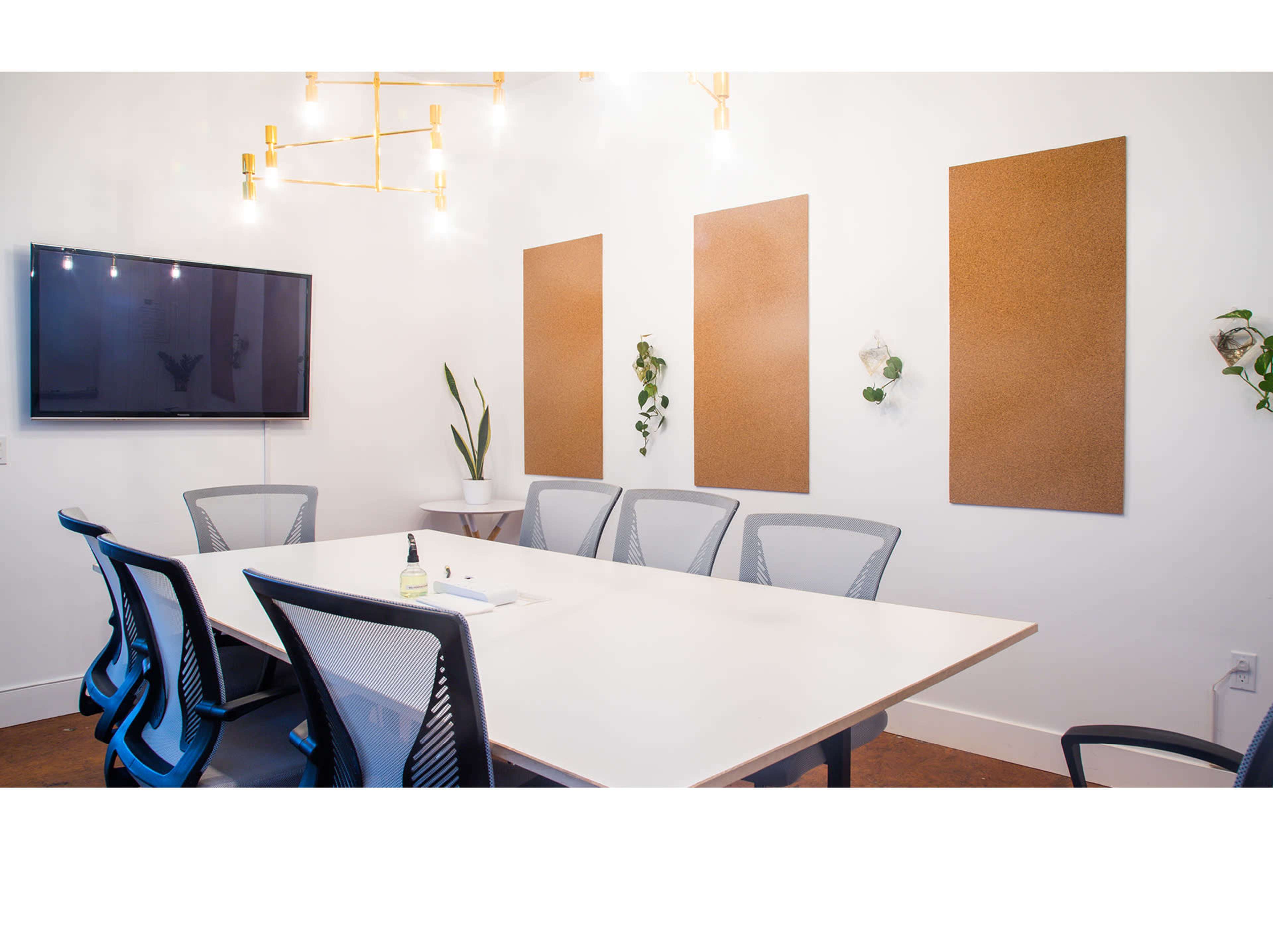 A bright conference room features a large white table surrounded by six black mesh chairs, with a television on the wall and three cork boards adorned with plants.