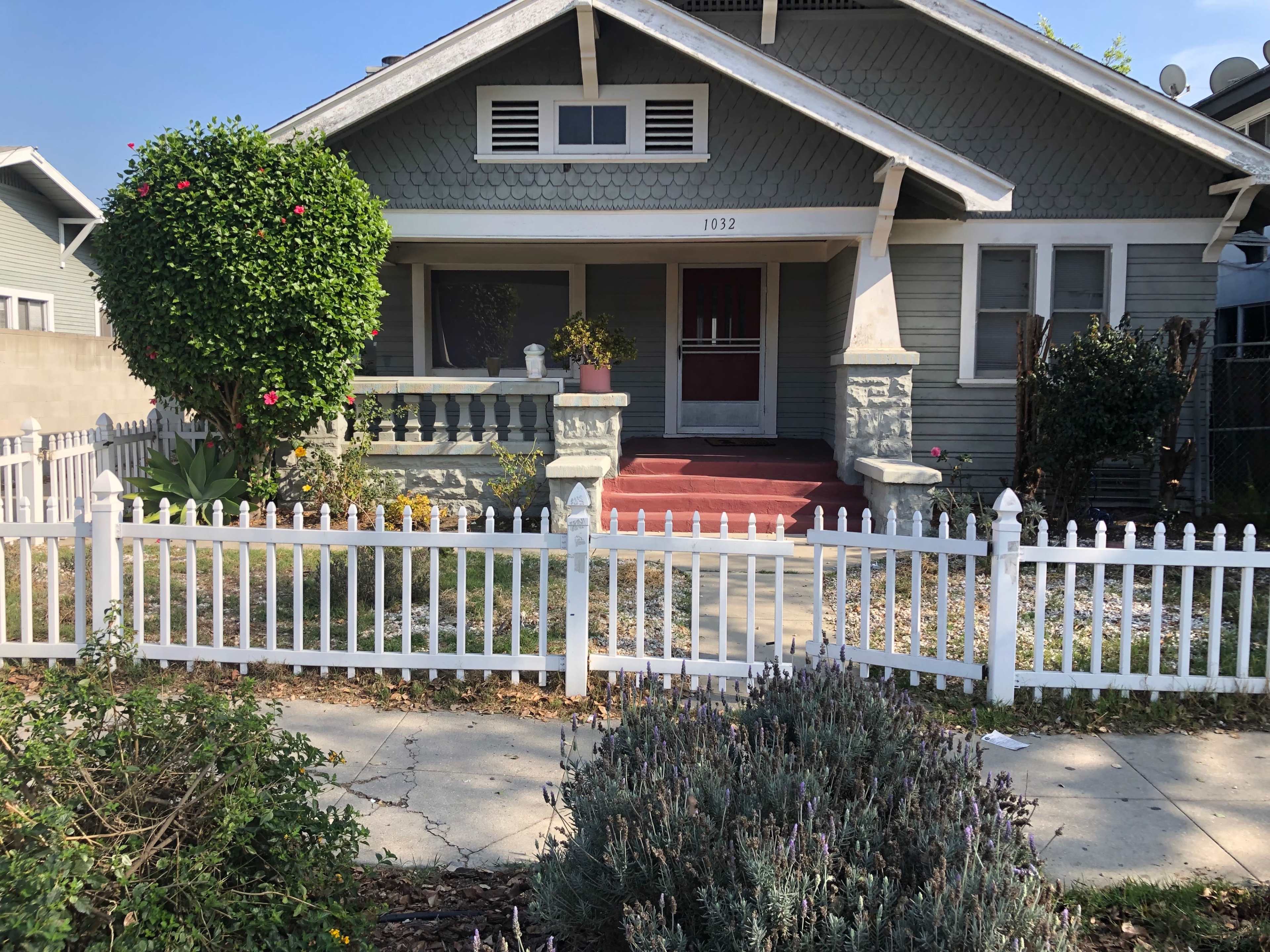 The image shows a green house with a porch, surrounded by a white picket fence and neatly landscaped with shrubs and flowers.