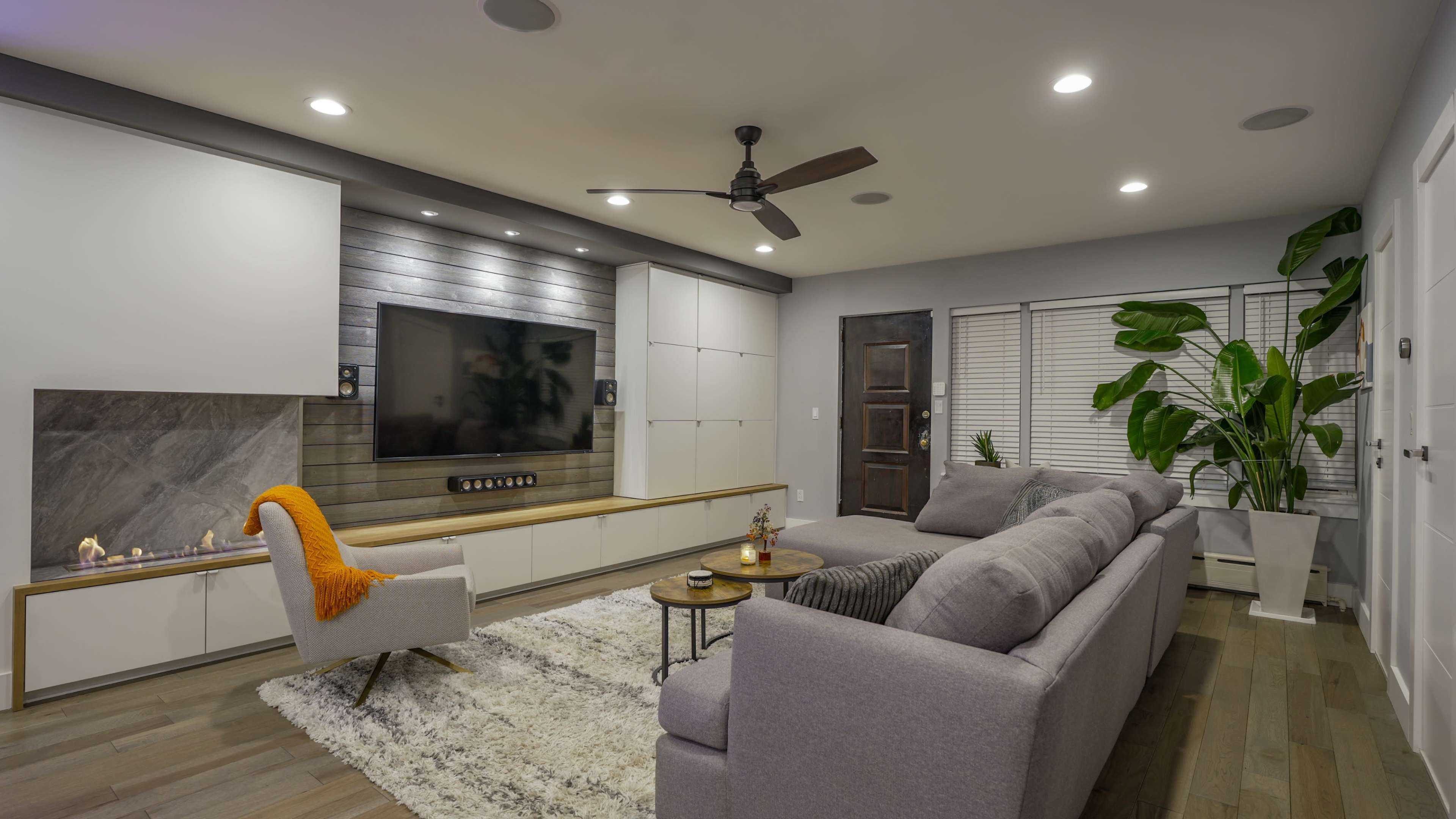 The image features a modern living room with a large sectional sofa, a wall-mounted television, and a decorative fireplace, complemented by plants and contemporary furnishings.