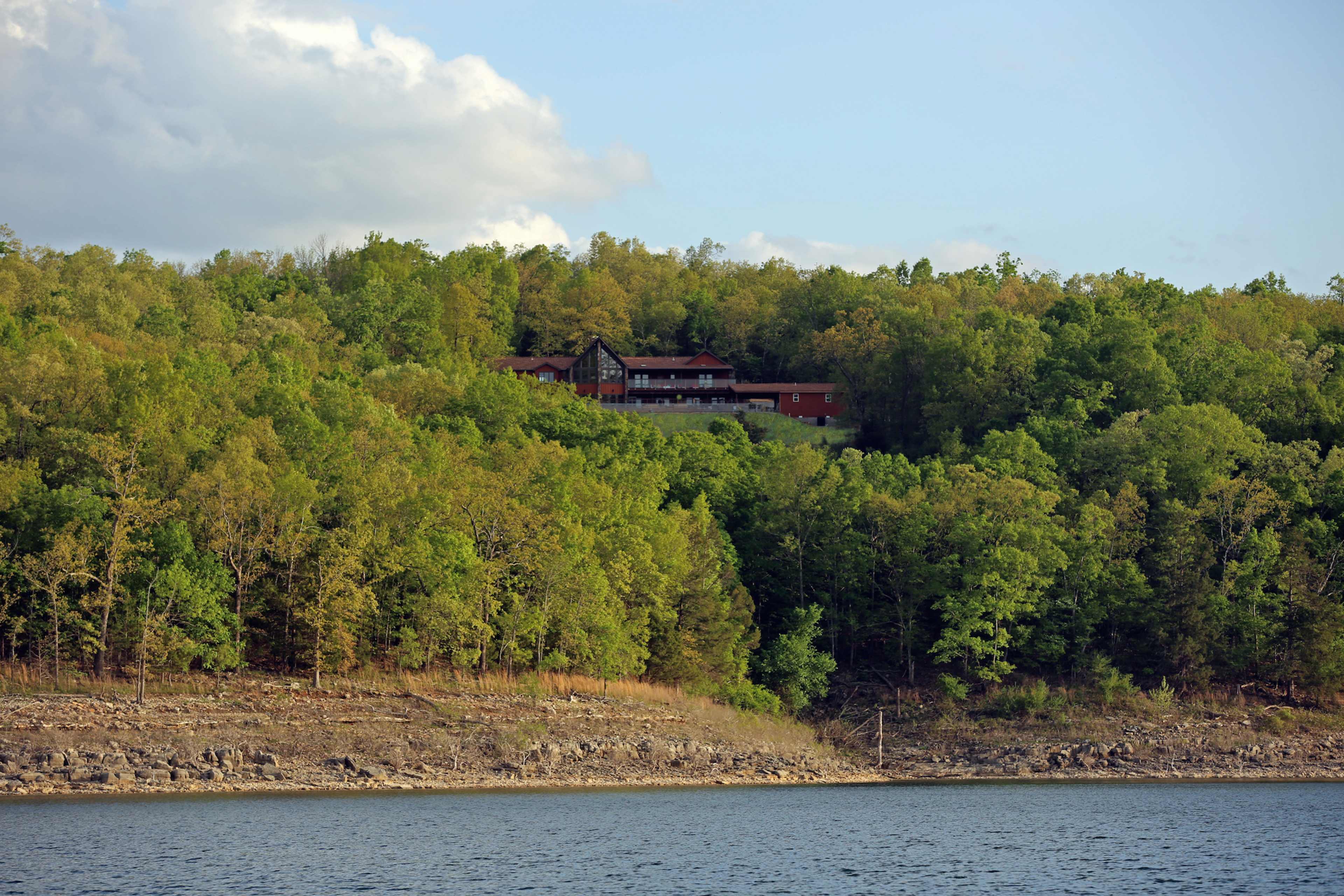 A large house sits atop a green, wooded hill overlooking a calm lake.